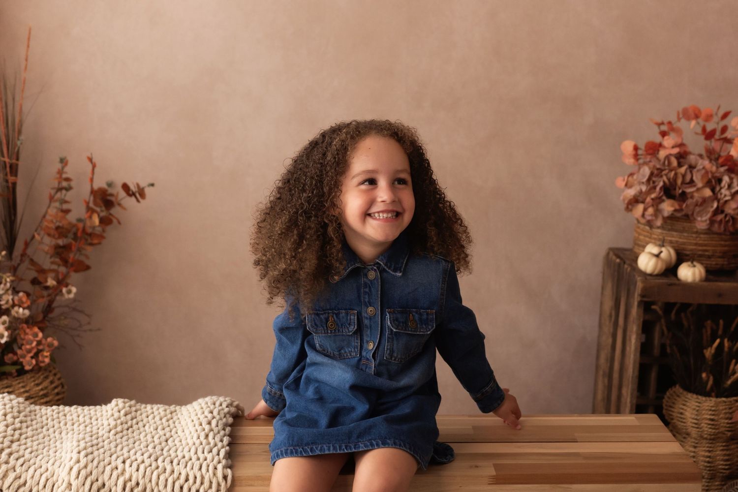 A smiling young girl with curly hair sits on a bench wearing a denim dress in an autumn-themed photo studio.