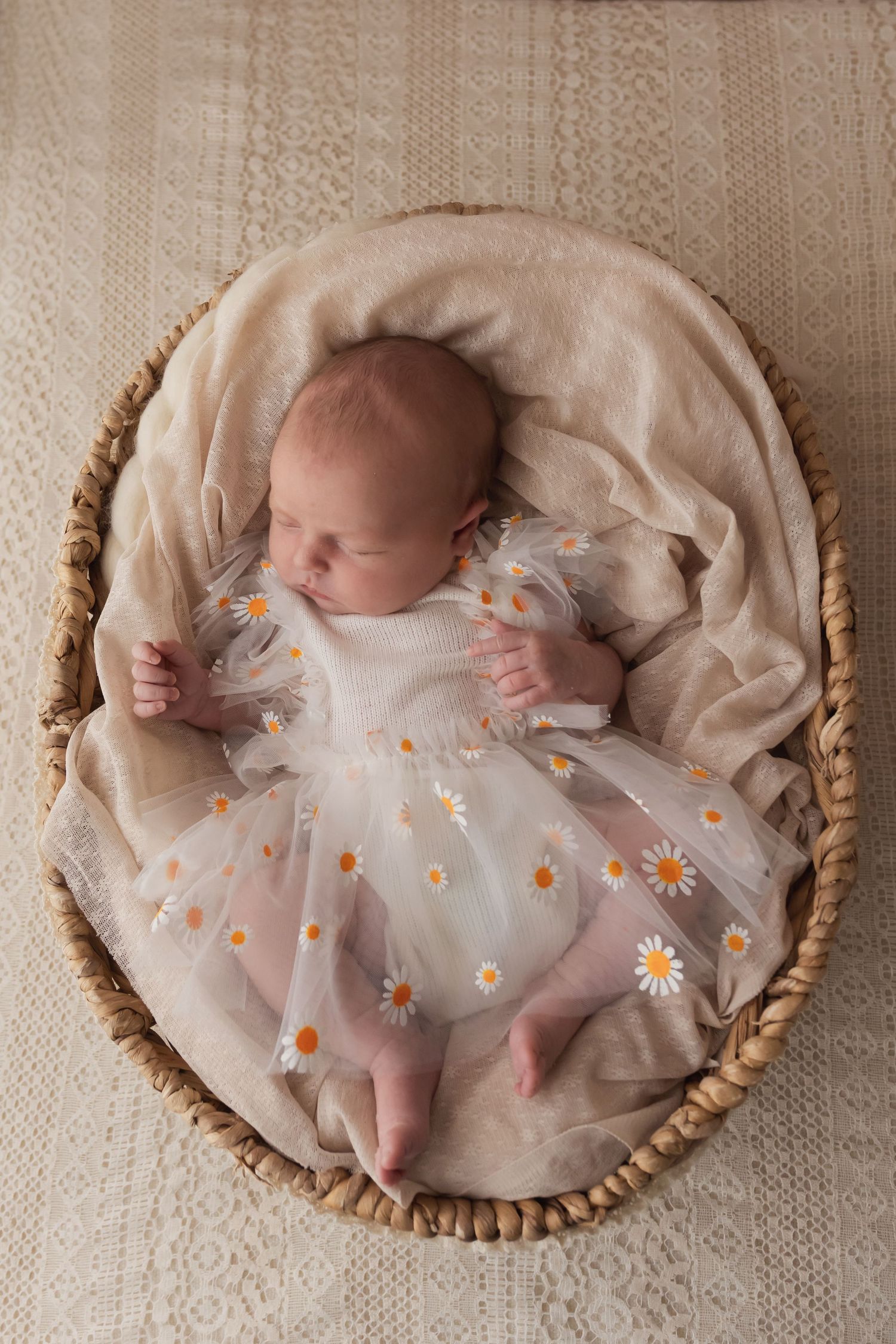 Sleeping newborn baby in daisy tulle dress lying in wicker basket on cream blanket.