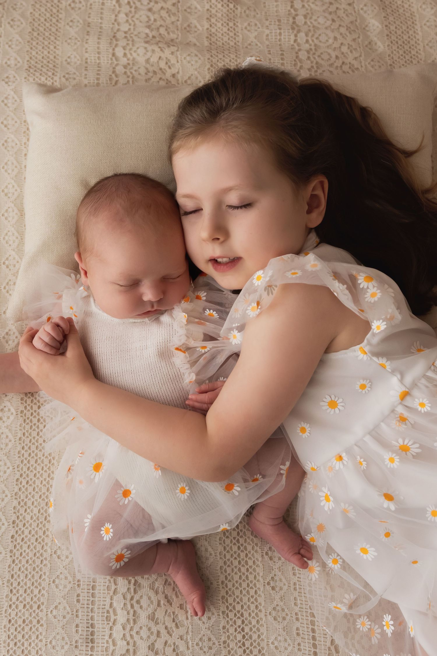Young girl in white daisy dress lying beside sleeping newborn sibling on cream blanket.