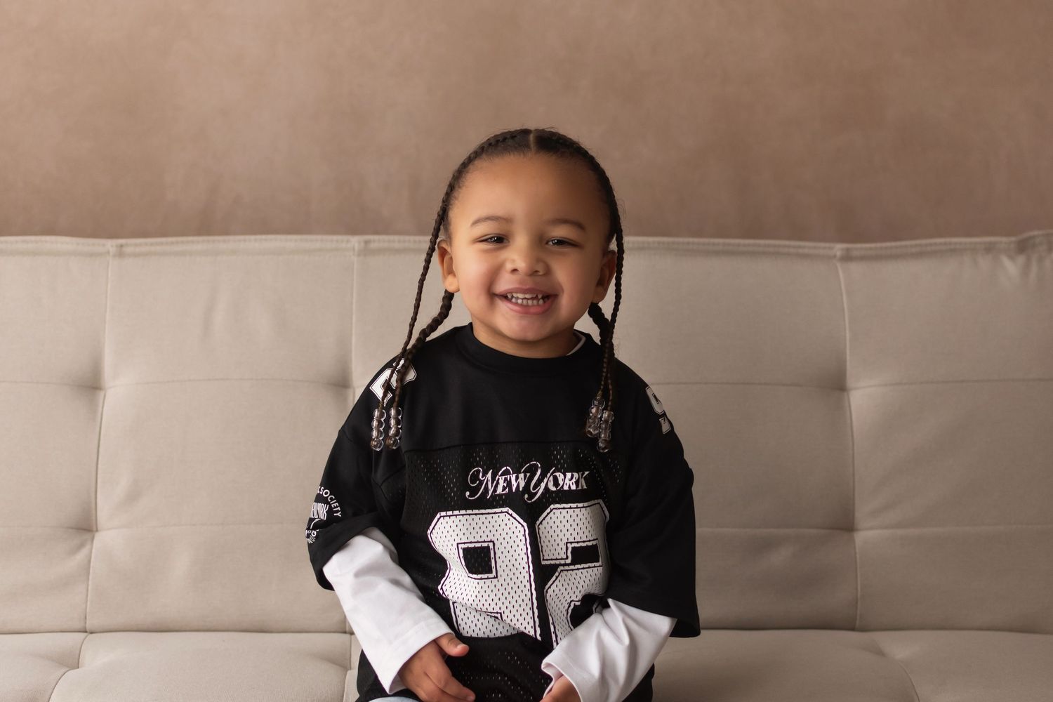 A happy young child with braids wearing a New York 92 jersey sits smiling on a gray couch in a casual portrait.