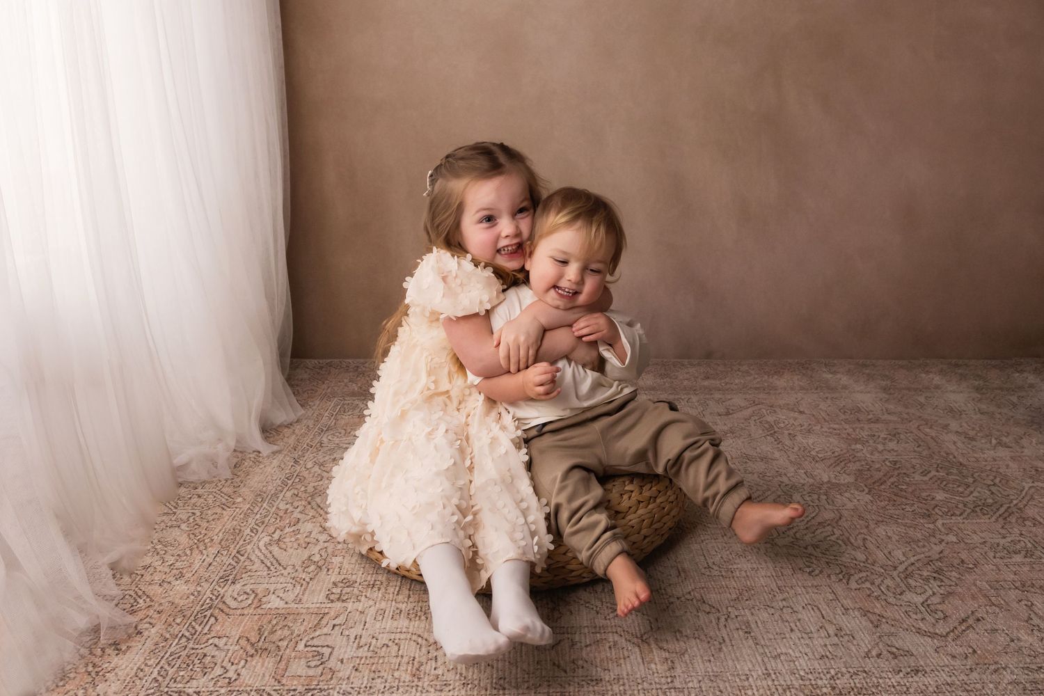 A young girl in a ruffled cream dress hugs her baby brother on a patterned rug in a warm studio setting.