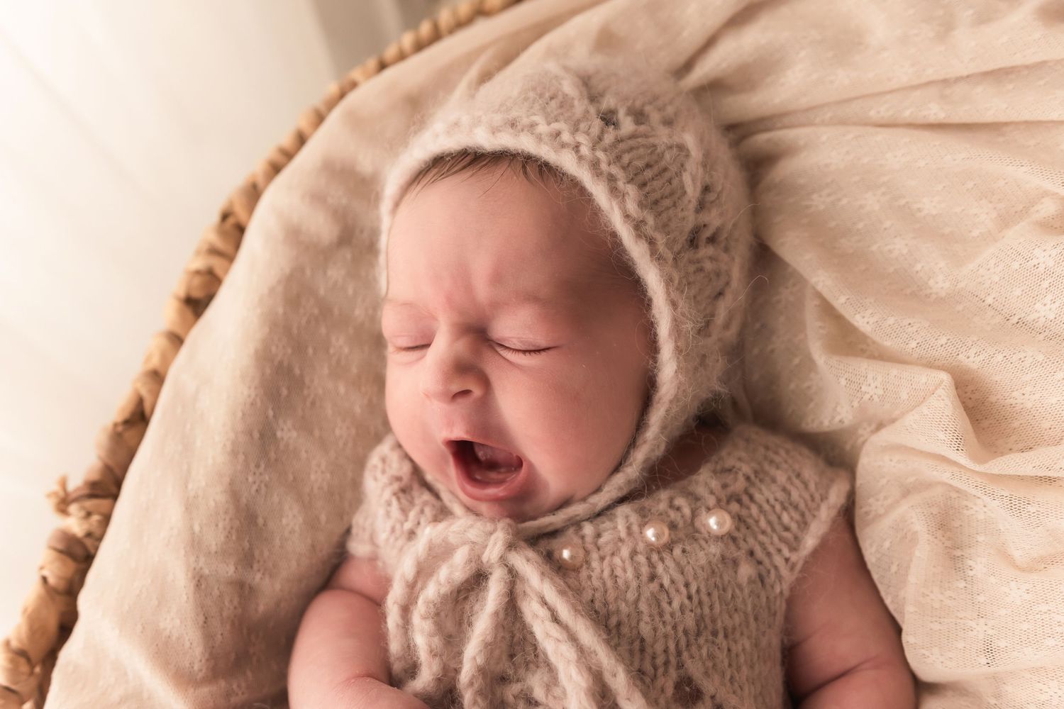 Newborn baby yawning in beige knit bonnet and pearl romper in wicker basket.