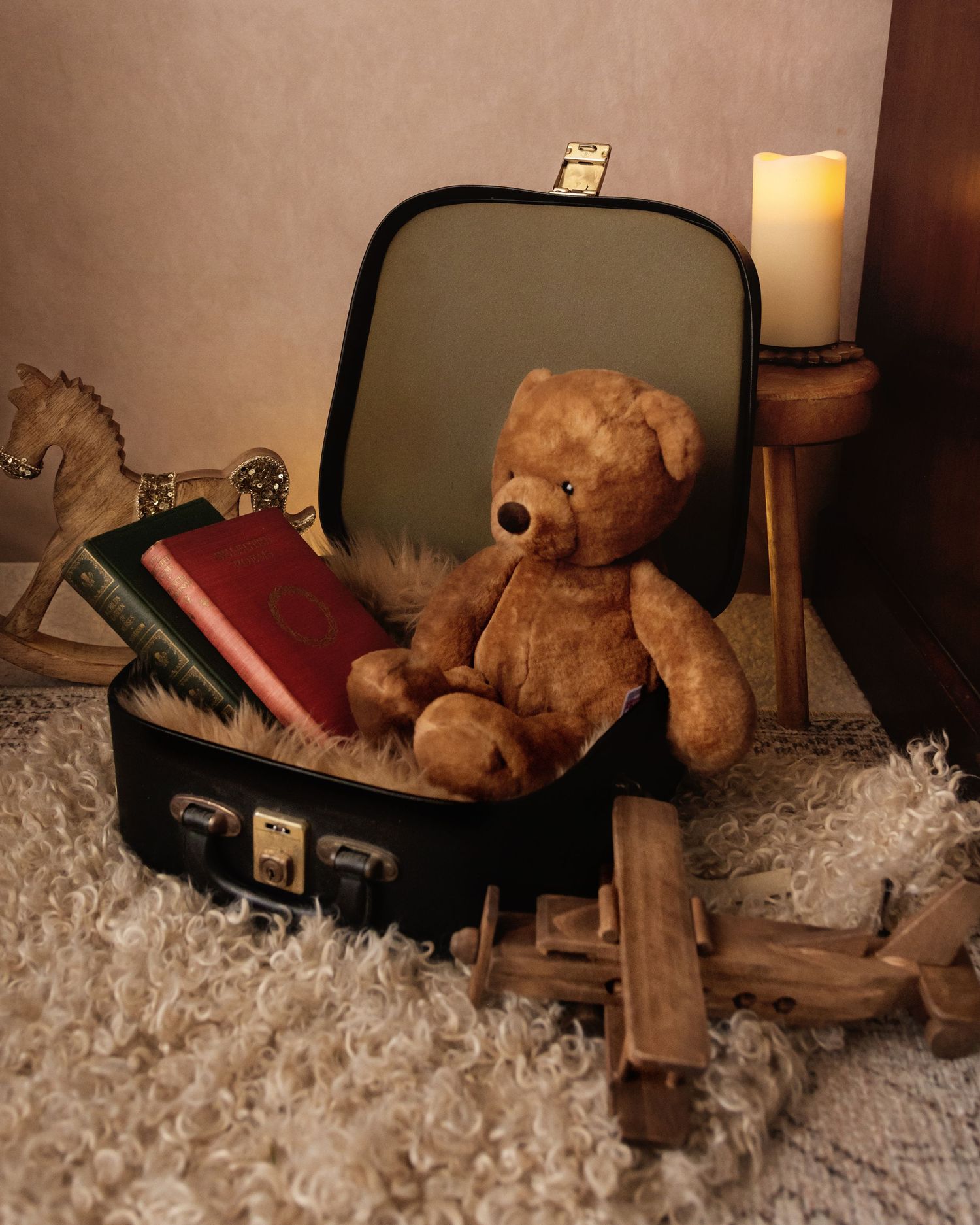 A vintage teddy bear sits in an open suitcase with old books, near a wooden toy horse and lit candle on a fluffy rug.