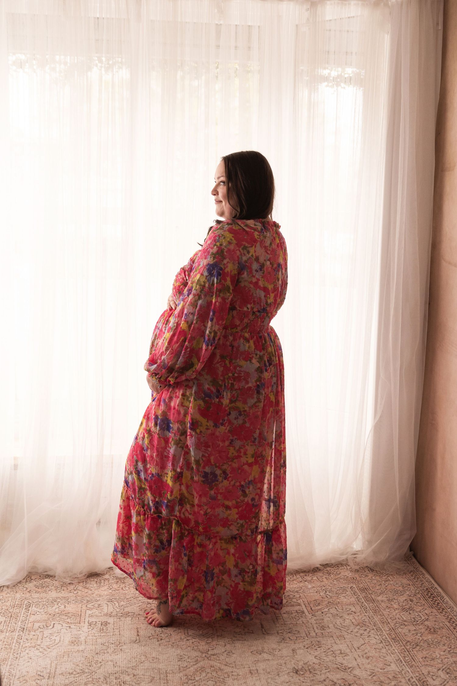 Pregnant woman in bright floral chiffon maxi dress standing before sheer white curtains, back view.