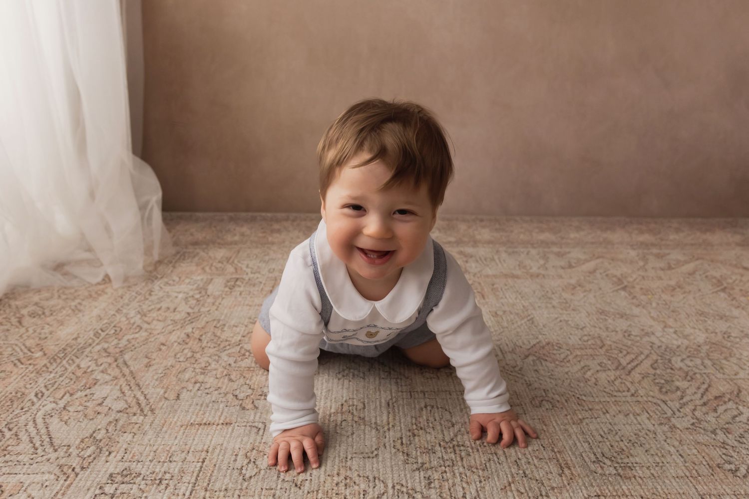 Cheerful baby boy crawling on patterned rug wearing white shirt and gray suspenders.