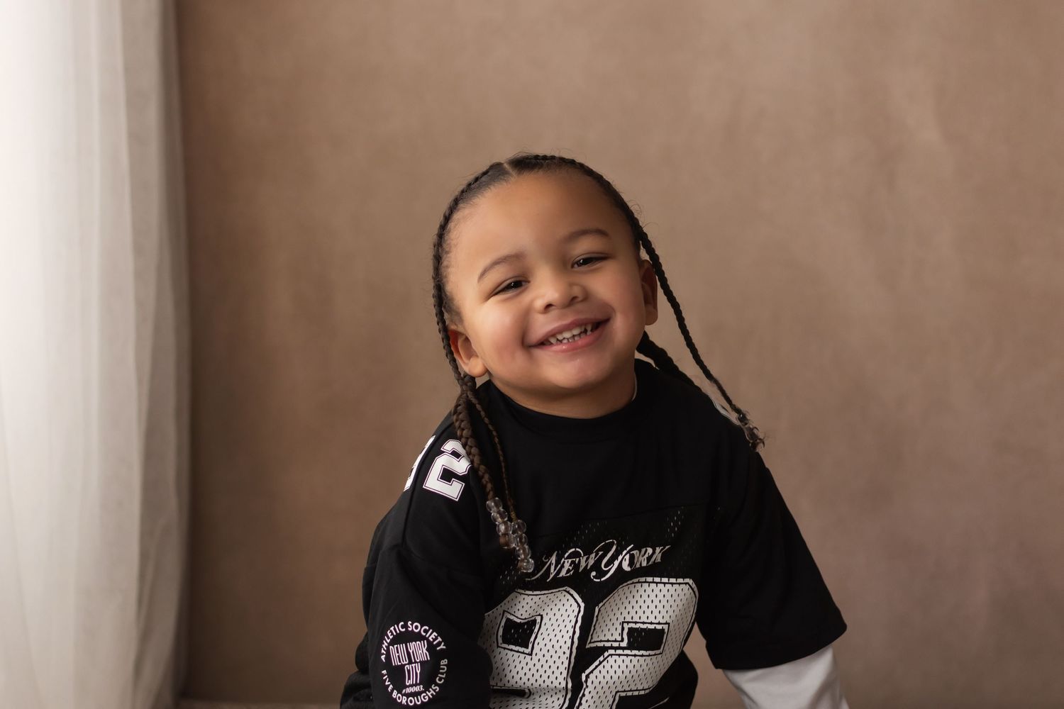 Happy young child with braids laughing in a black New York 92 jersey against a brown backdrop.