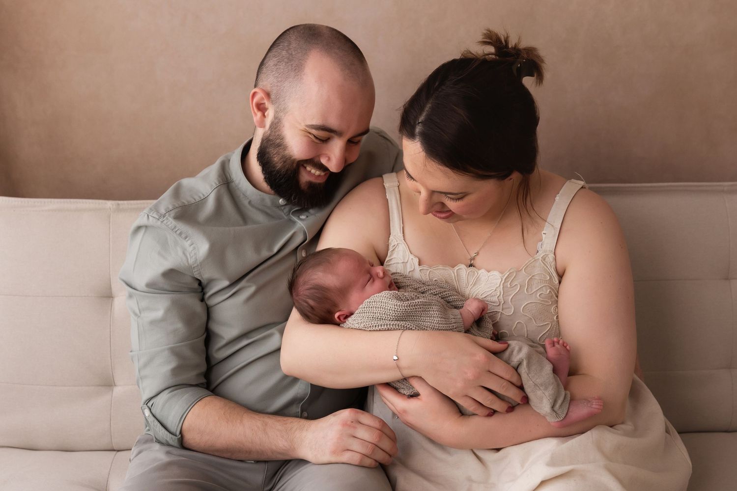 New parents sitting on couch gazing lovingly at their sleeping newborn baby.