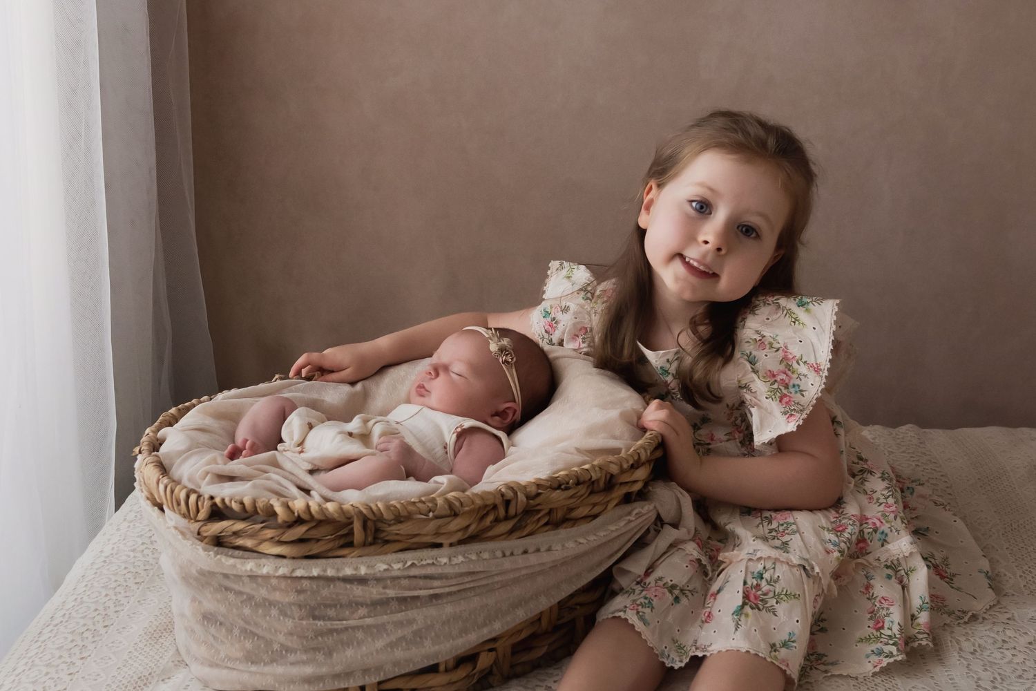Young girl in floral dress posing with sleeping newborn in wicker basket.