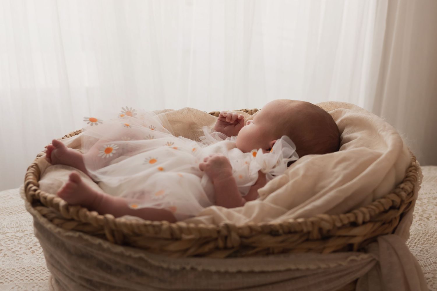 Newborn baby in floral tulle dress sleeping peacefully in wicker basket near window.