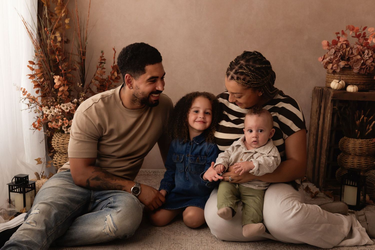 Happy family of four sitting together, parents smiling at young daughter while mother holds baby in cozy studio setting.