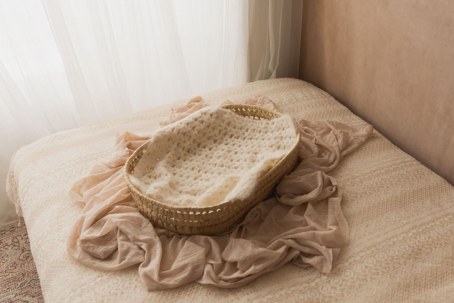 Woven rattan basket with crochet lining on beige fabric draped over a soft neutral cushion near sheer curtains.