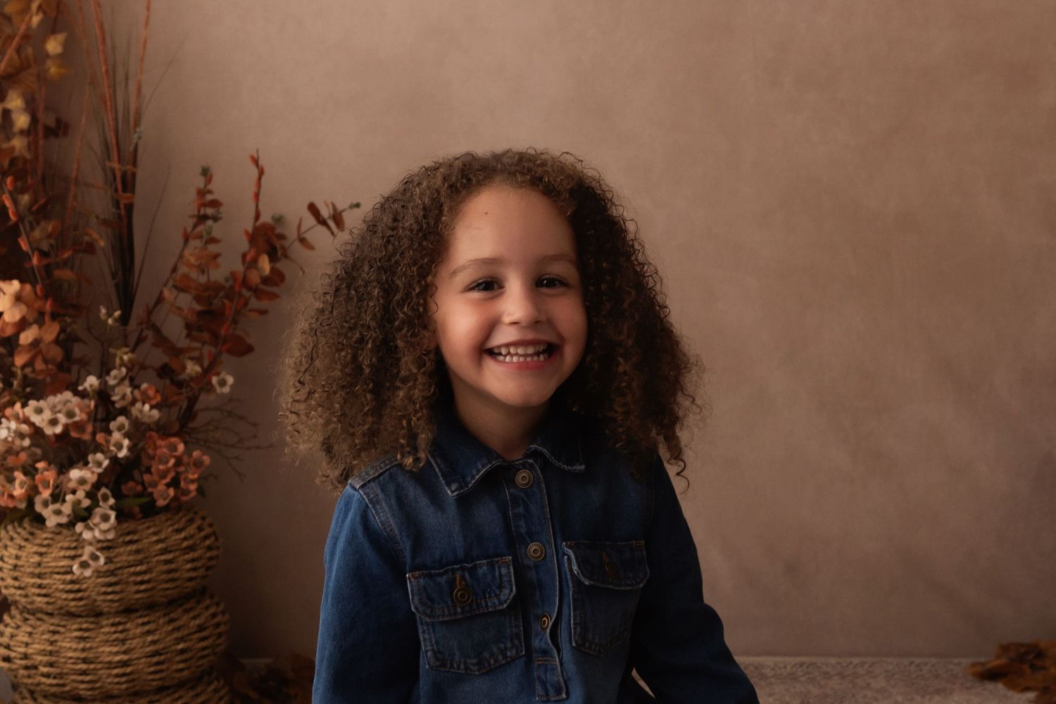 A young girl with curly hair smiles brightly in a denim jacket, posed before a warm beige backdrop with autumn florals.