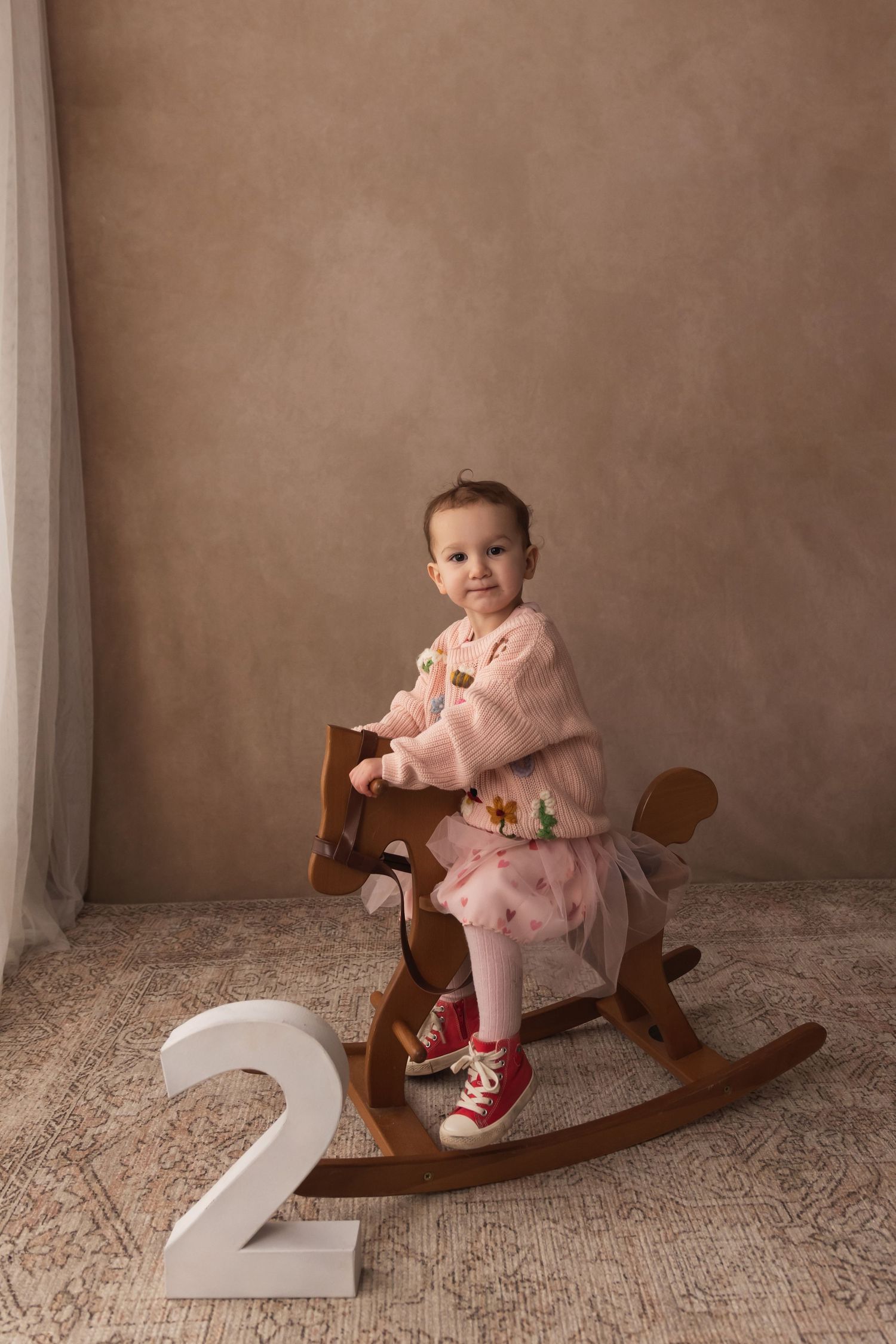 Toddler girl in pink outfit riding wooden rocking horse beside number 2 for second birthday photos.