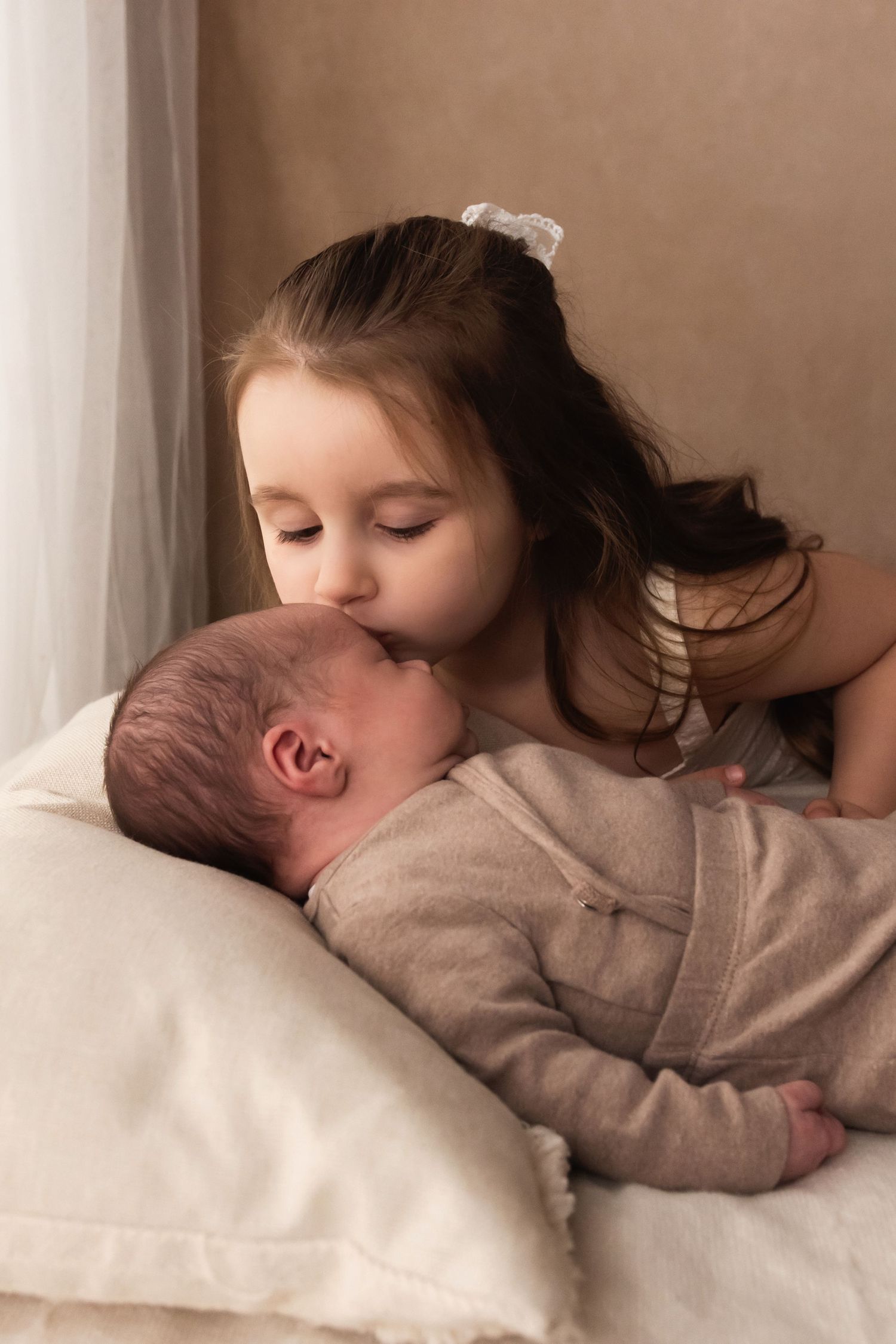 Older sister gently kissing sleeping newborn baby on forehead on white pillow.