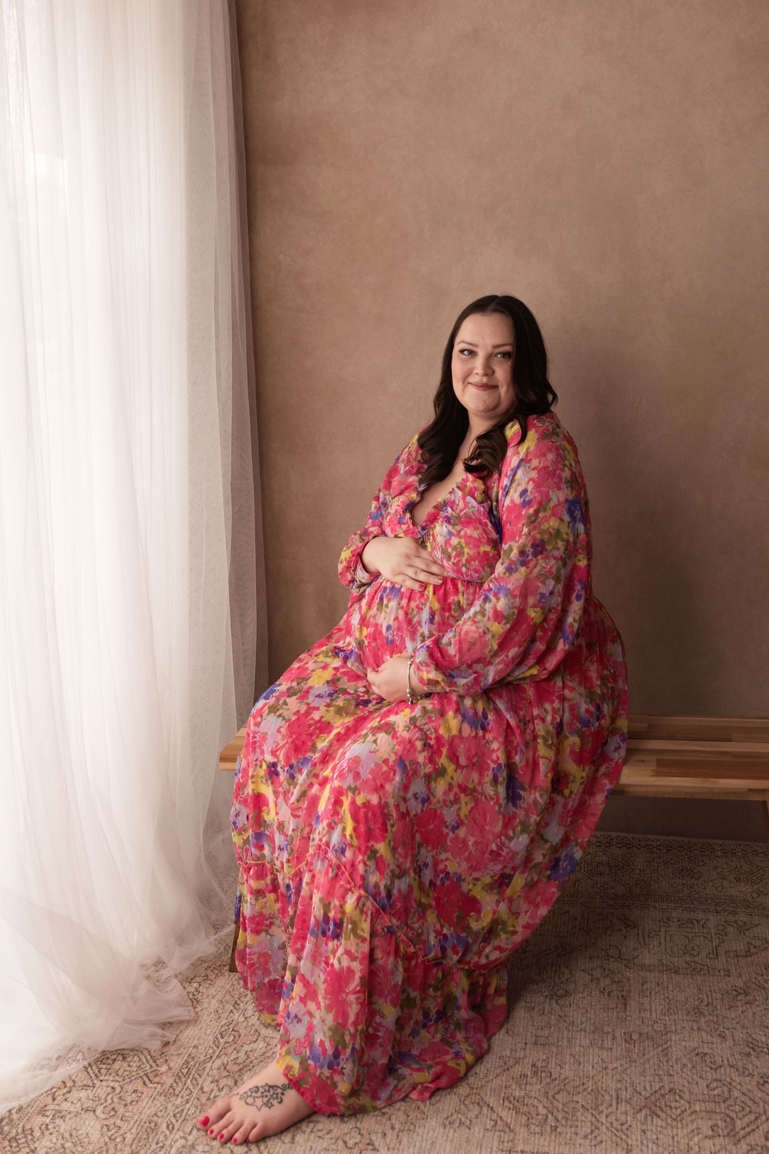 Smiling pregnant woman in a colorful pink floral maxi dress sitting gracefully beside a sheer curtain.