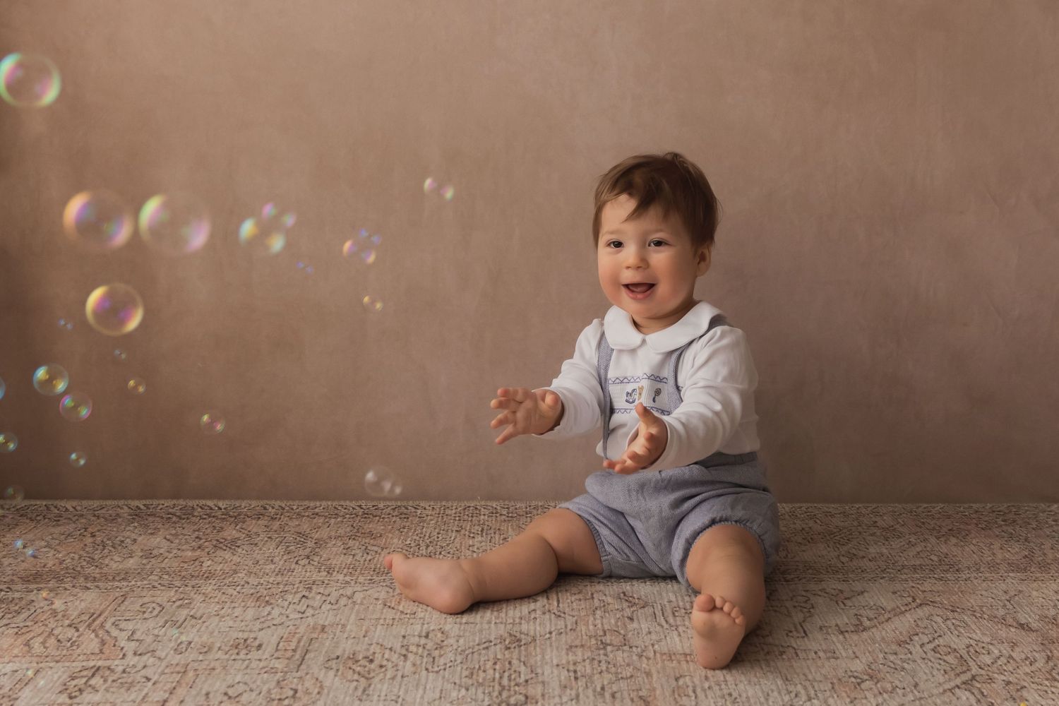 Delighted baby boy in gray overalls reaches for floating soap bubbles during first birthday photoshoot.