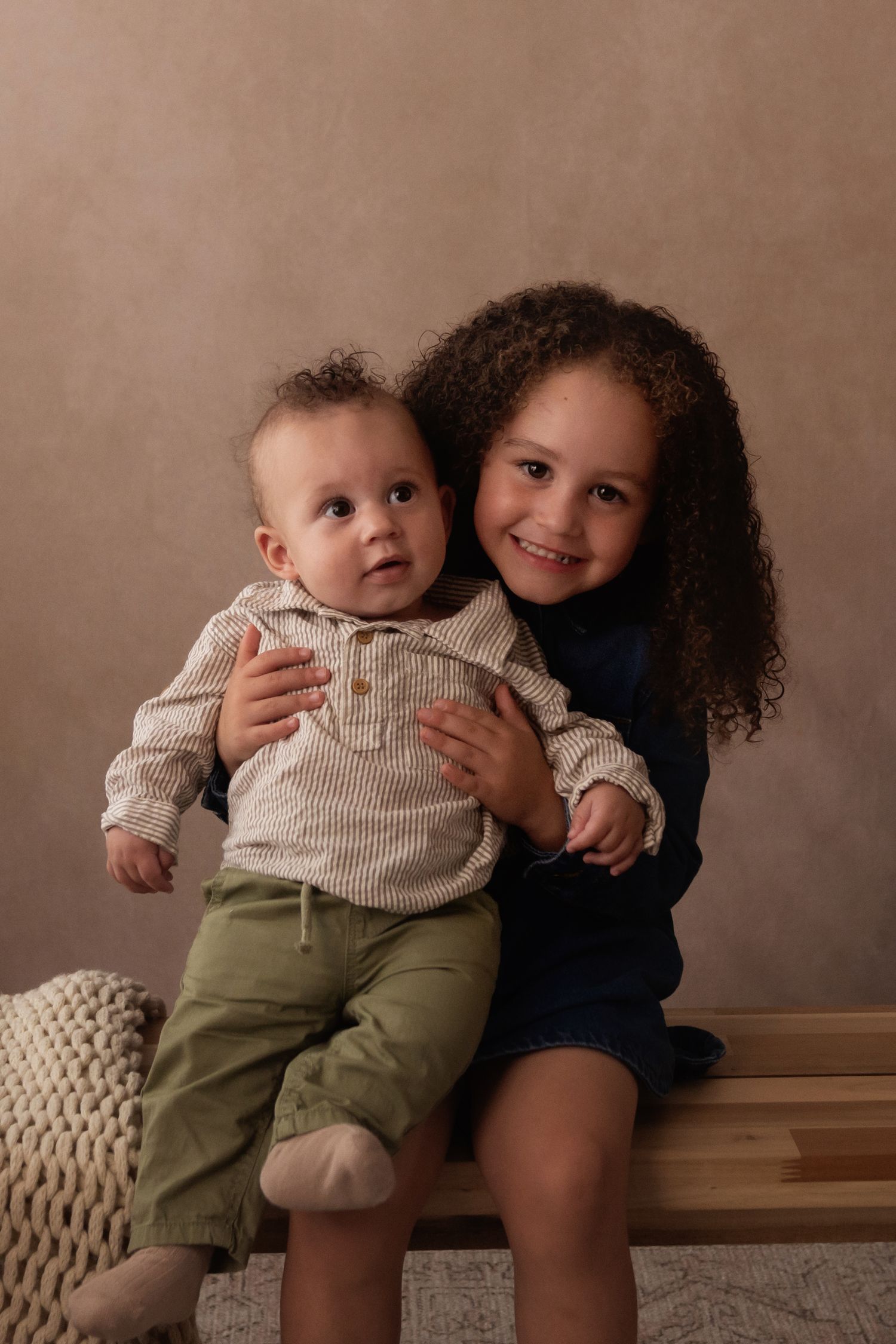 A smiling girl with curly hair hugs a baby boy in a studio portrait setting.