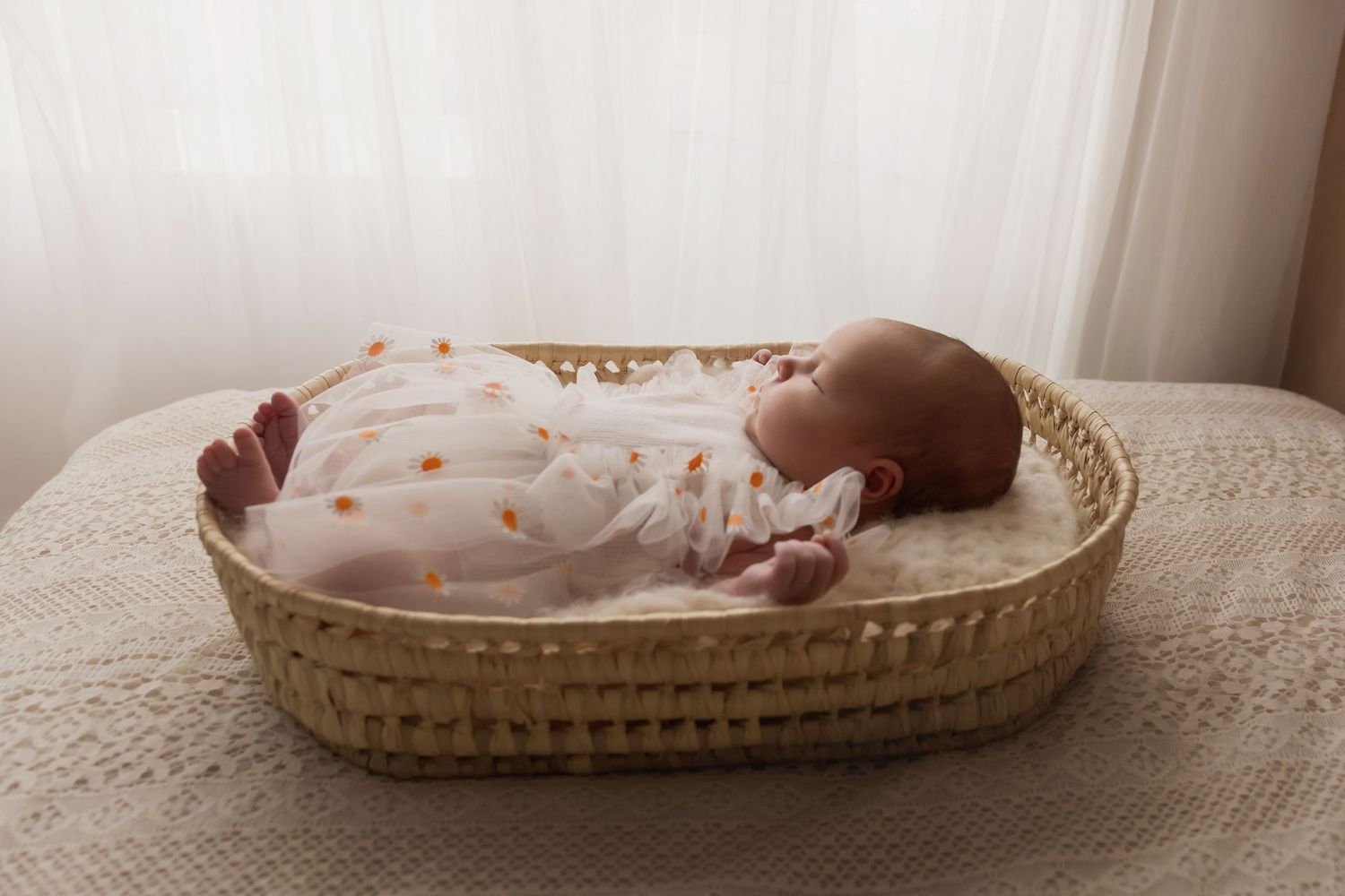 Sleeping newborn baby in white floral outfit lying in a wicker Moses basket by window.