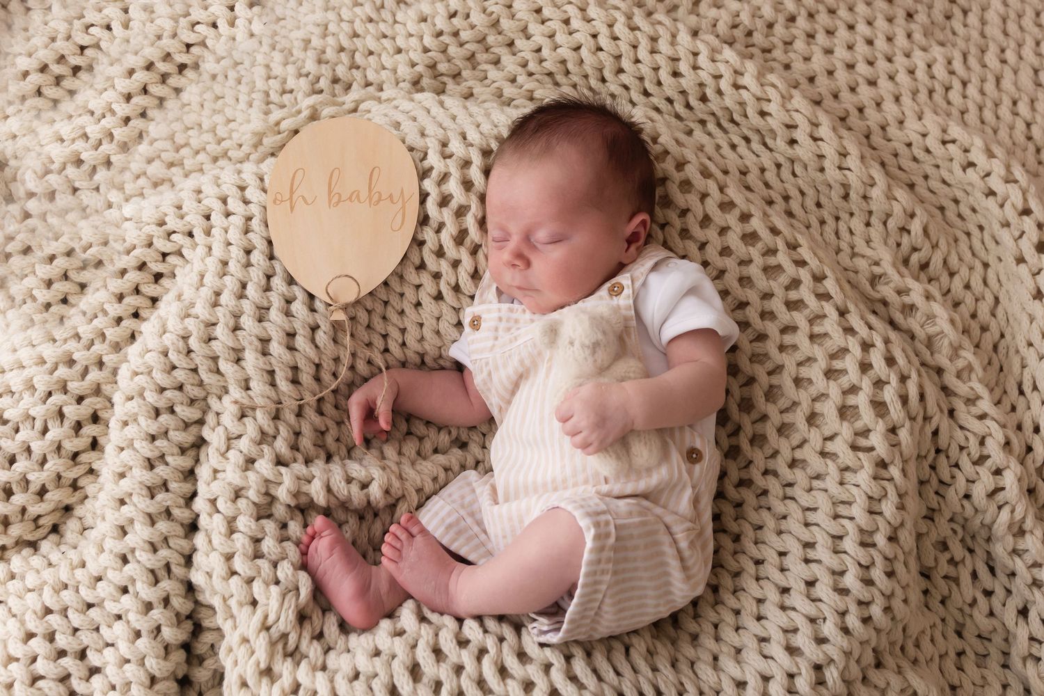 Sleeping newborn baby in cream knit outfit beside a wooden name disc on chunky knit blanket.