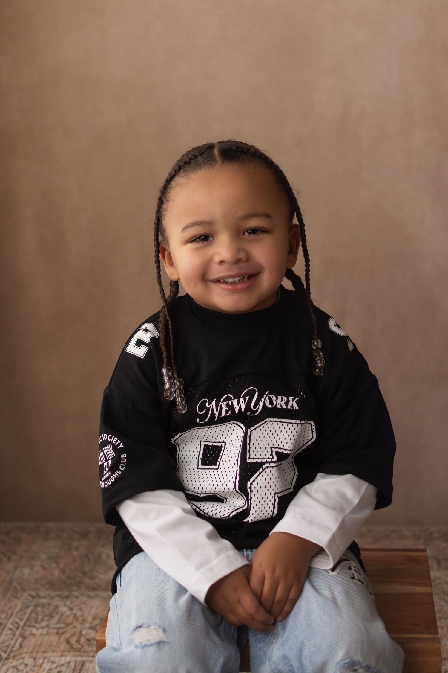 A smiling young child with braids wearing a New York 92 jersey and ripped jeans sits in a warm studio setting.