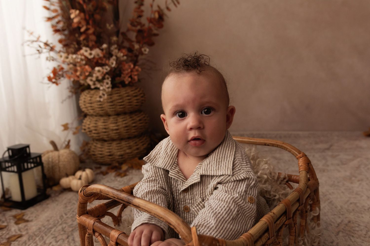 Adorable baby boy sitting in a wicker basket with fall autumn decor, pumpkins, and dried flowers in a warm studio setting.