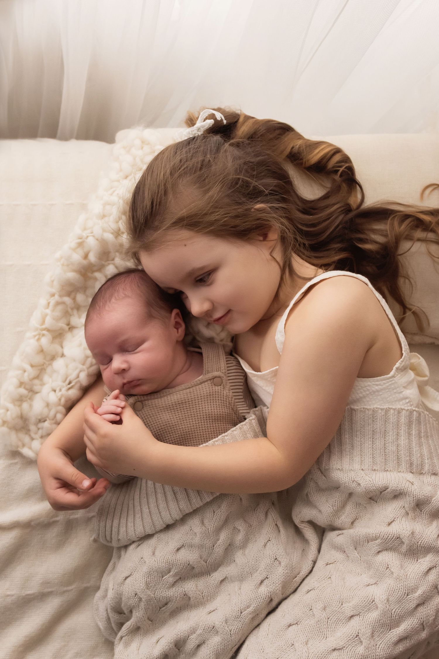 Big sister lying down cuddling and holding sleeping newborn baby in knit outfit on blanket.