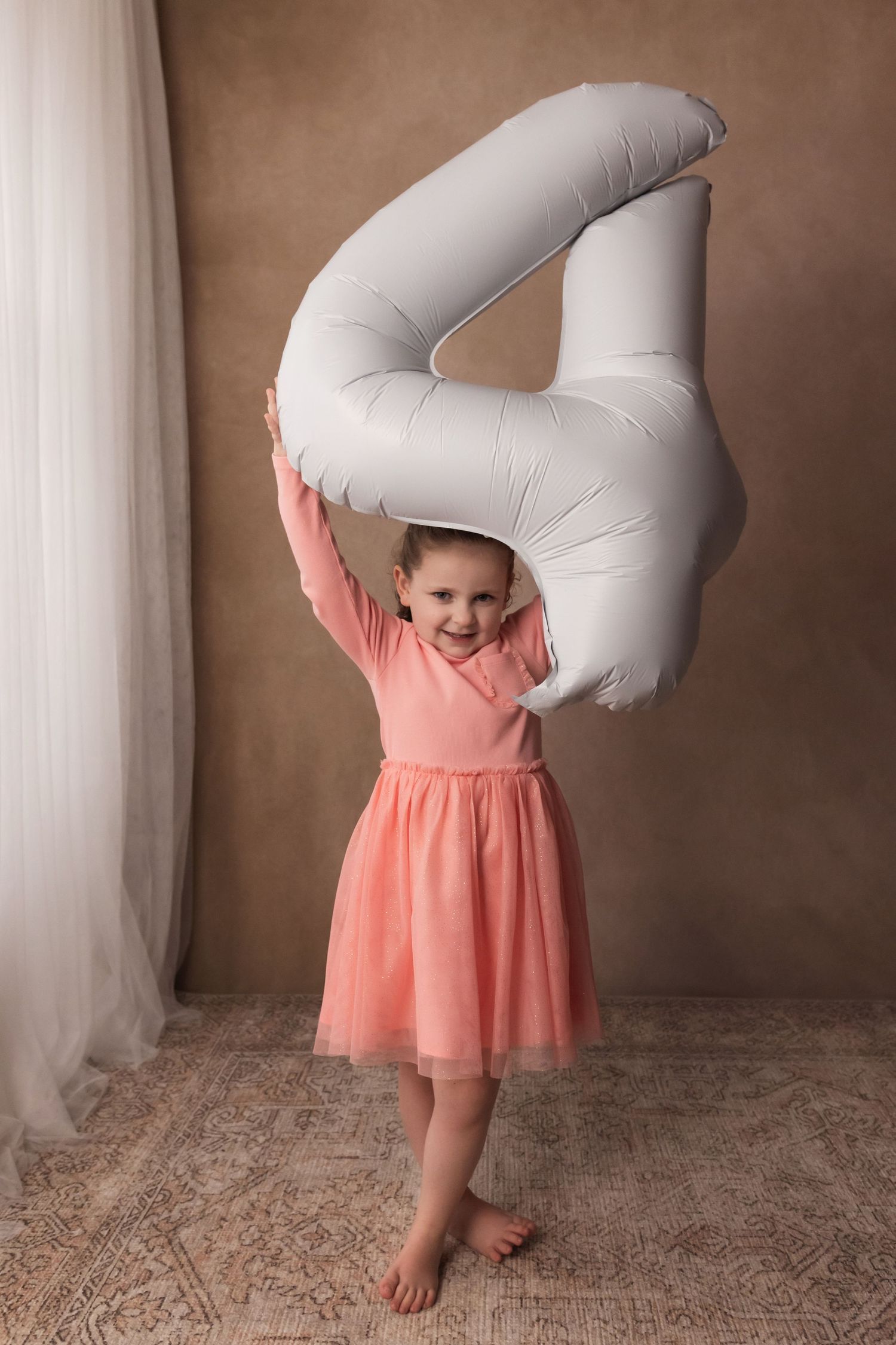 Young girl in pink dress holding large silver number 4 balloon overhead for birthday portraits.