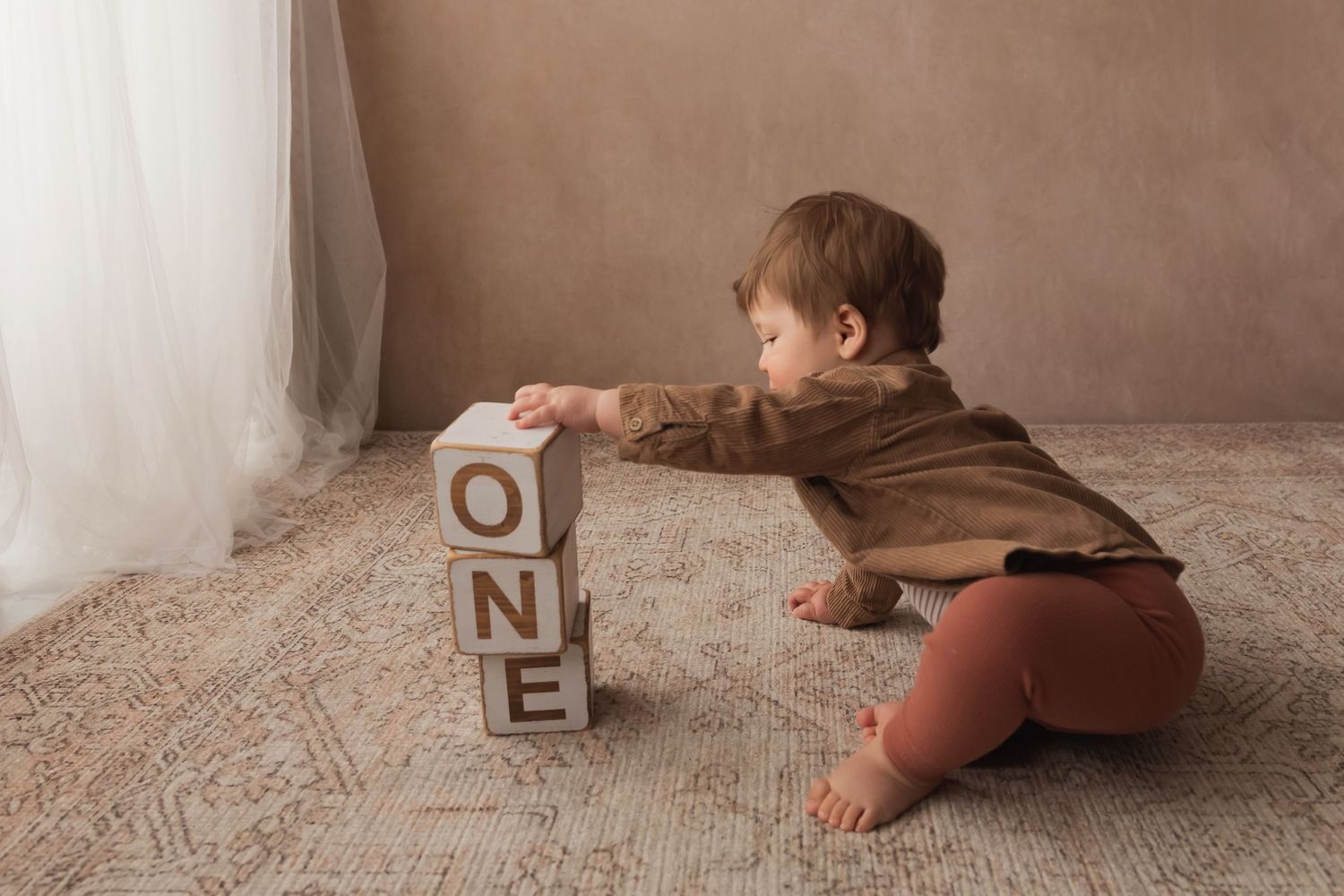 Baby in brown outfit stacking wooden ONE letter blocks on a patterned rug.