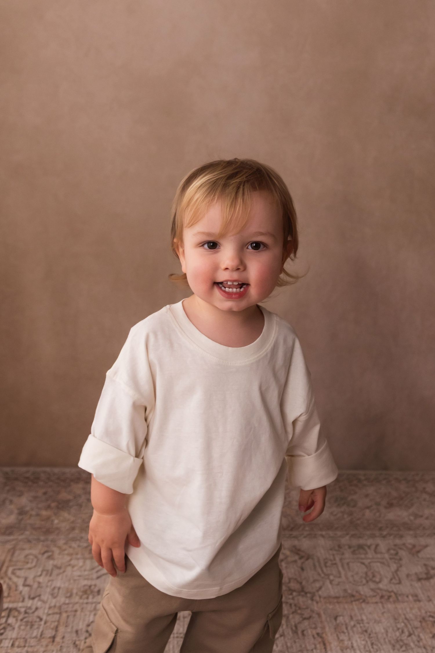 An adorable toddler in an oversized white long-sleeve shirt smiles at the camera in a warm studio setting.