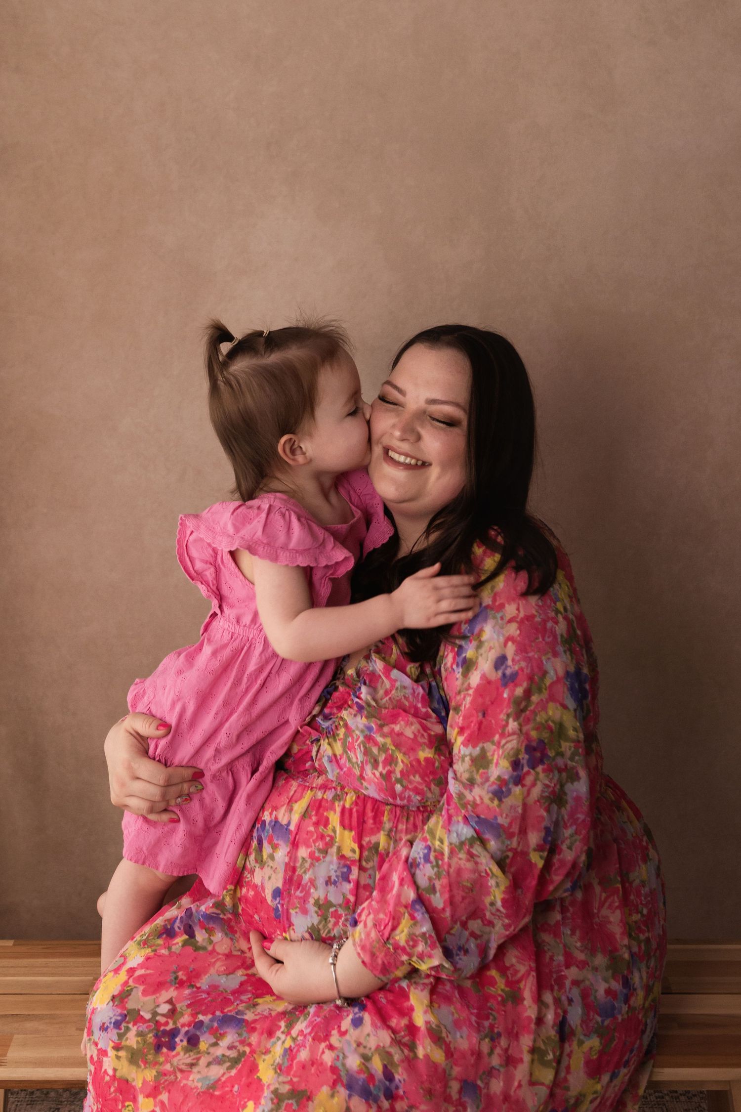 Young girl in pink dress kissing smiling pregnant mother in floral dress on the cheek.