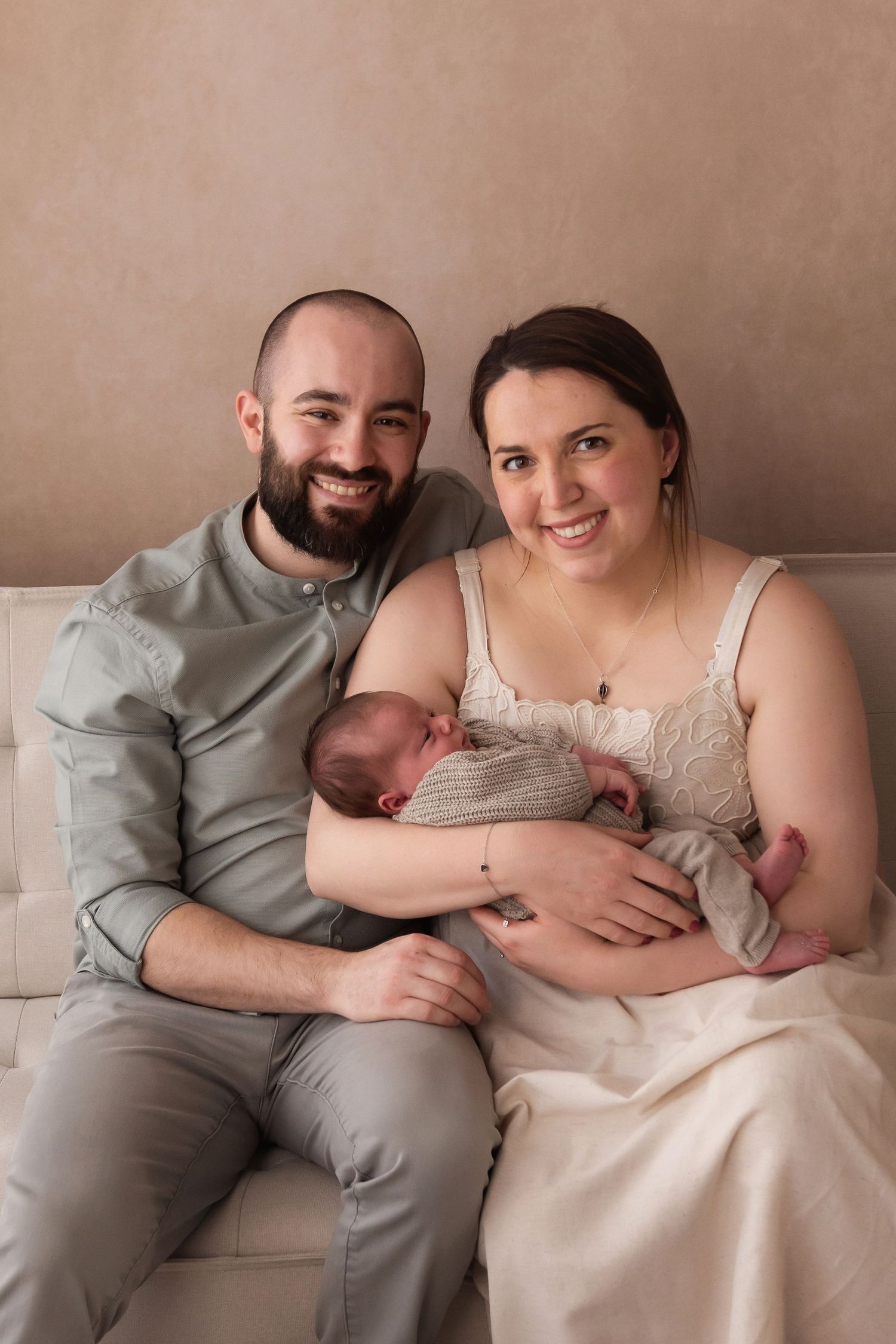 Smiling couple sitting on a couch holding their newborn baby, dressed in soft neutral tones for a family portrait.