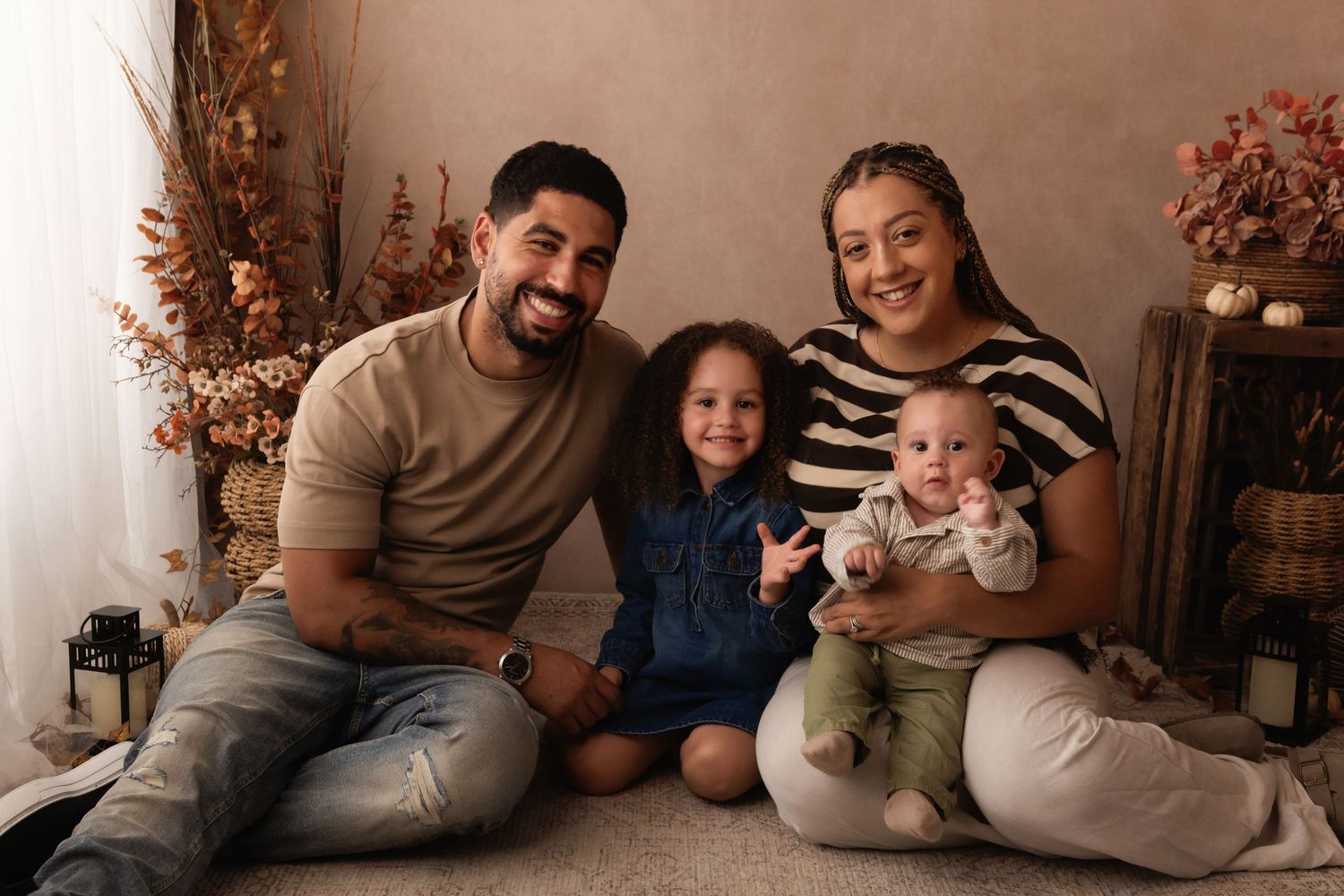 Happy family of four posing for a portrait, with two parents, a toddler girl, and a baby boy in a warm studio setting.