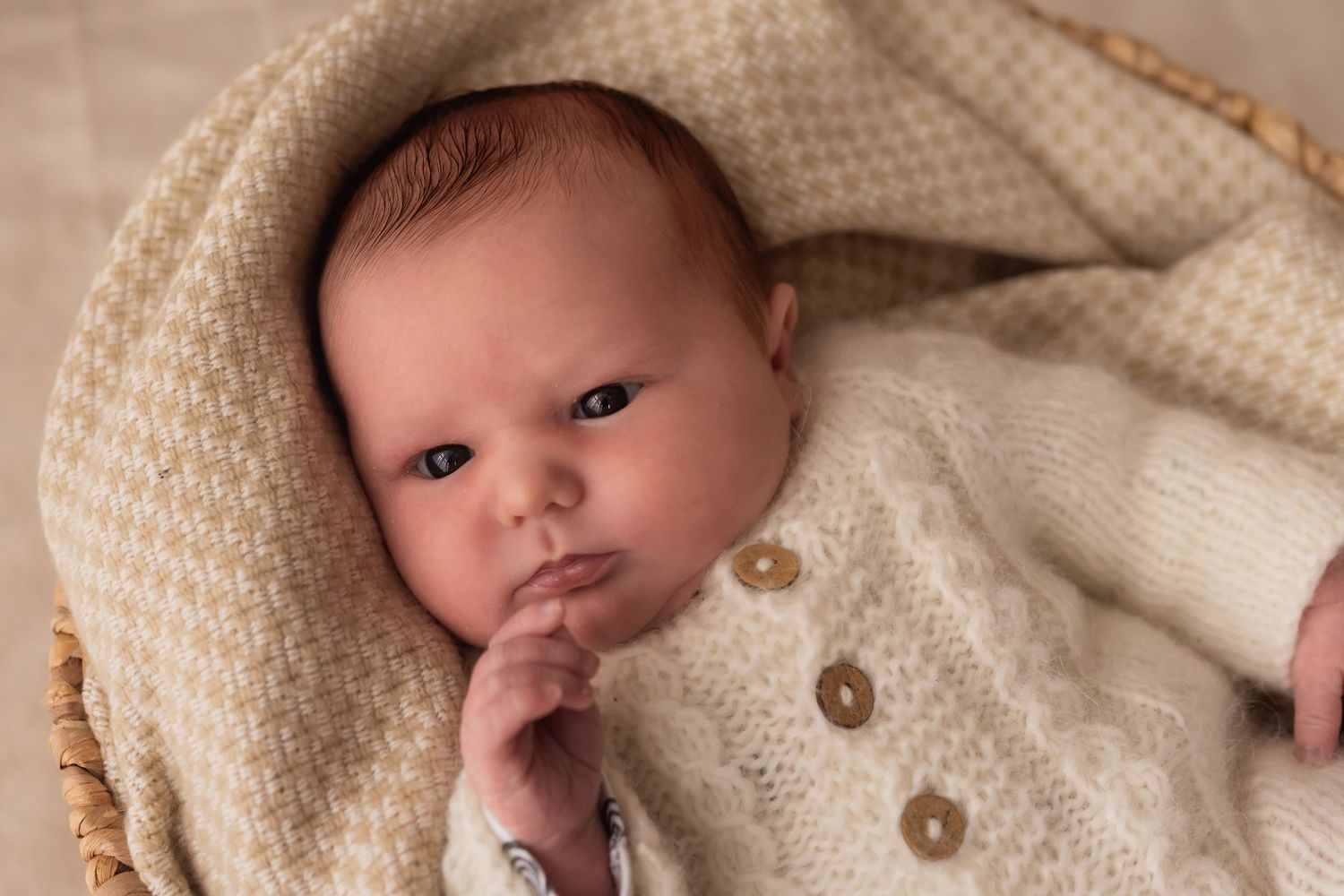 Alert newborn baby with finger near mouth wrapped in cream knit blanket in wicker basket.