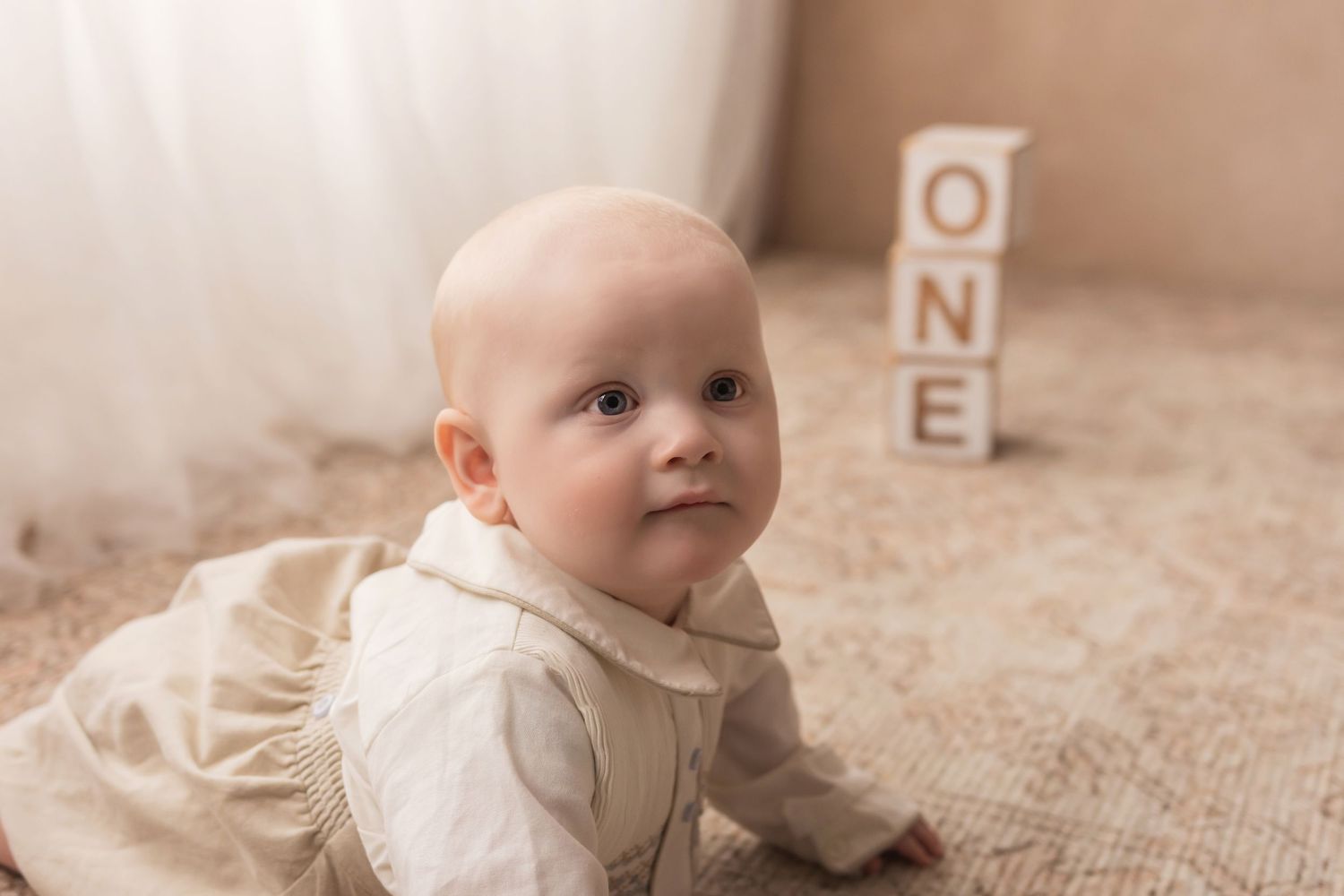 Curious baby in cream romper crawls near wooden ONE blocks on a textured beige rug.