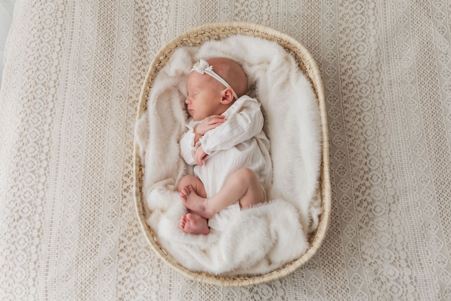 Sleeping newborn baby in a white outfit resting in a woven oval basket lined with soft white fur.