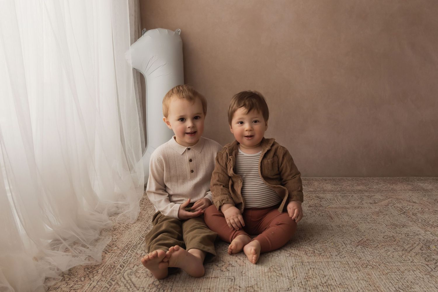 Two young brothers in neutral outfits sitting together on a rug beside a number one balloon.