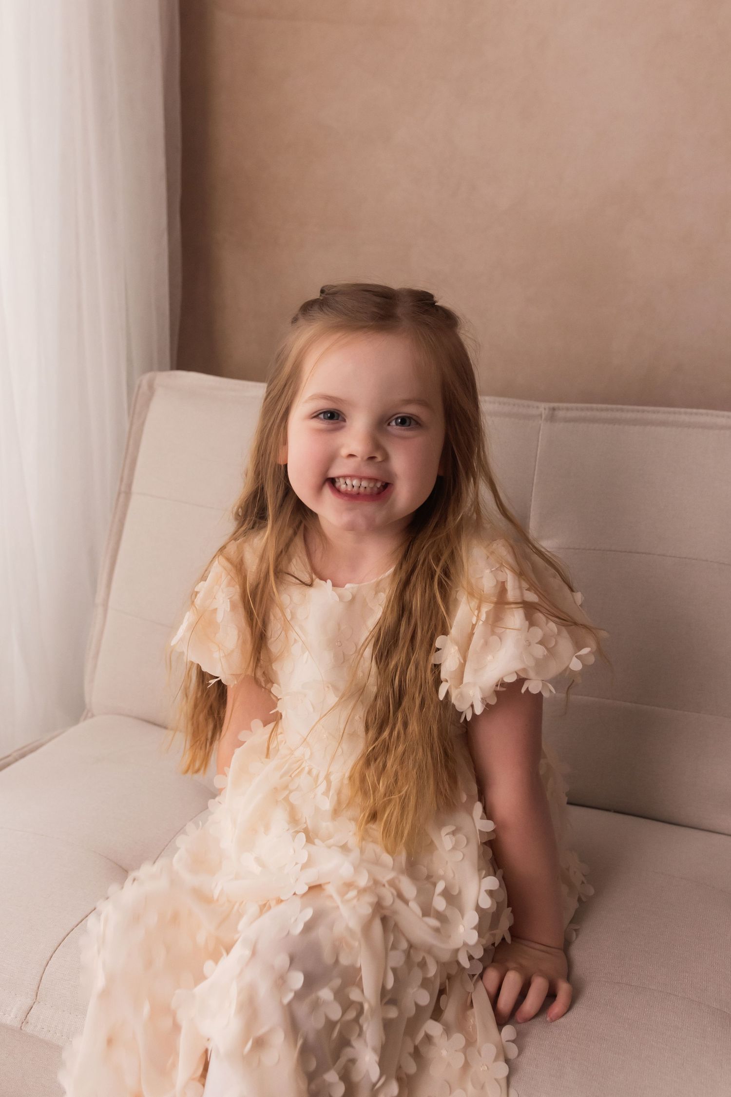A smiling young girl with long blonde hair wearing a cream floral ruffle dress, sitting on a white sofa.