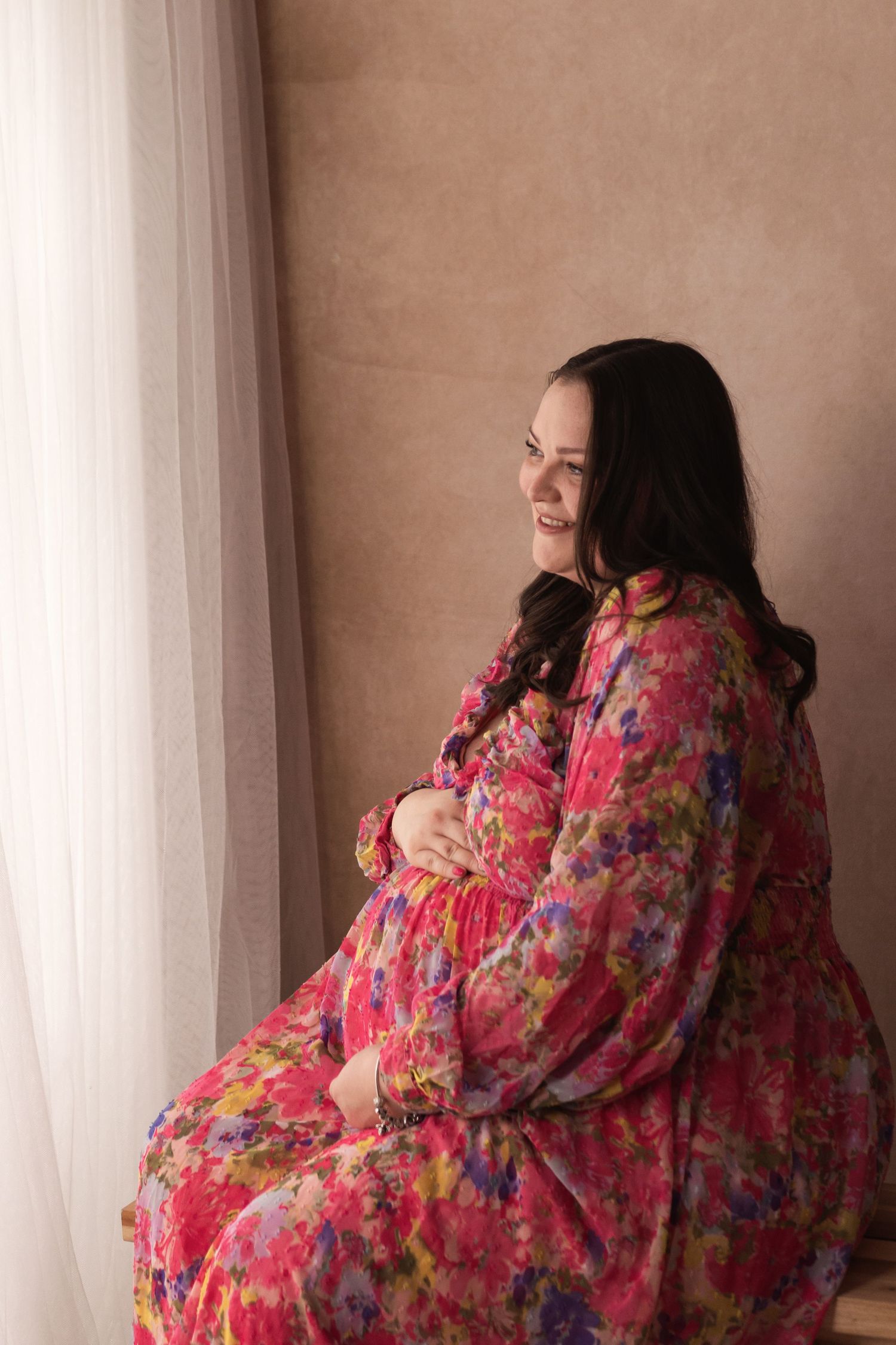 Smiling pregnant woman in colorful floral dress sitting and holding her baby bump near a window.