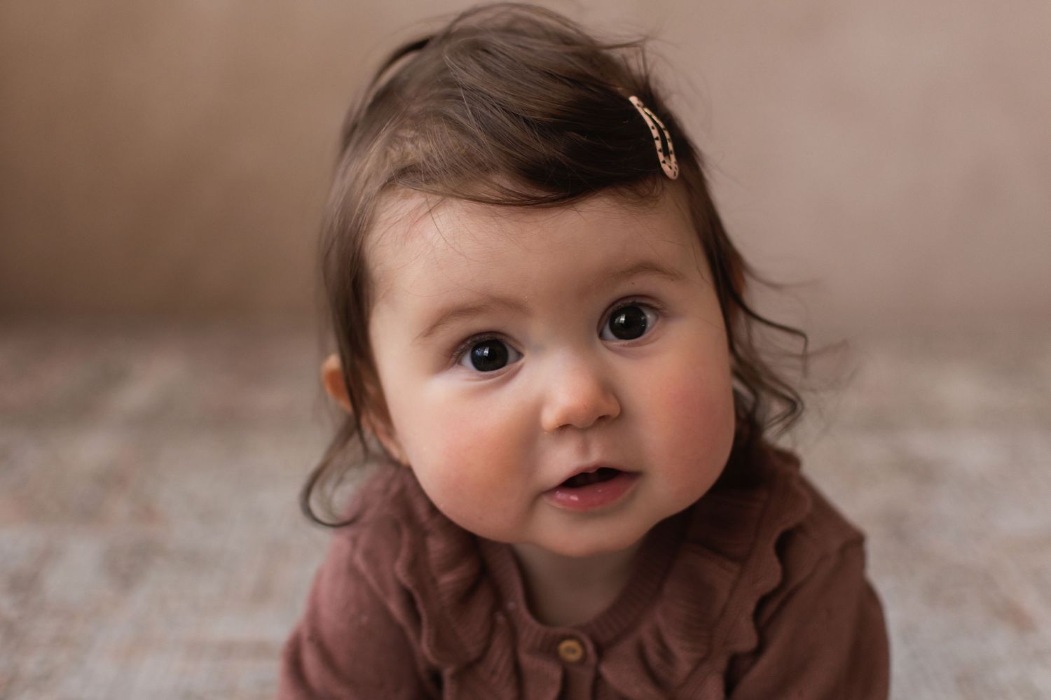 A wide-eyed baby girl in a brown ruffle romper looks up curiously in a professional portrait.