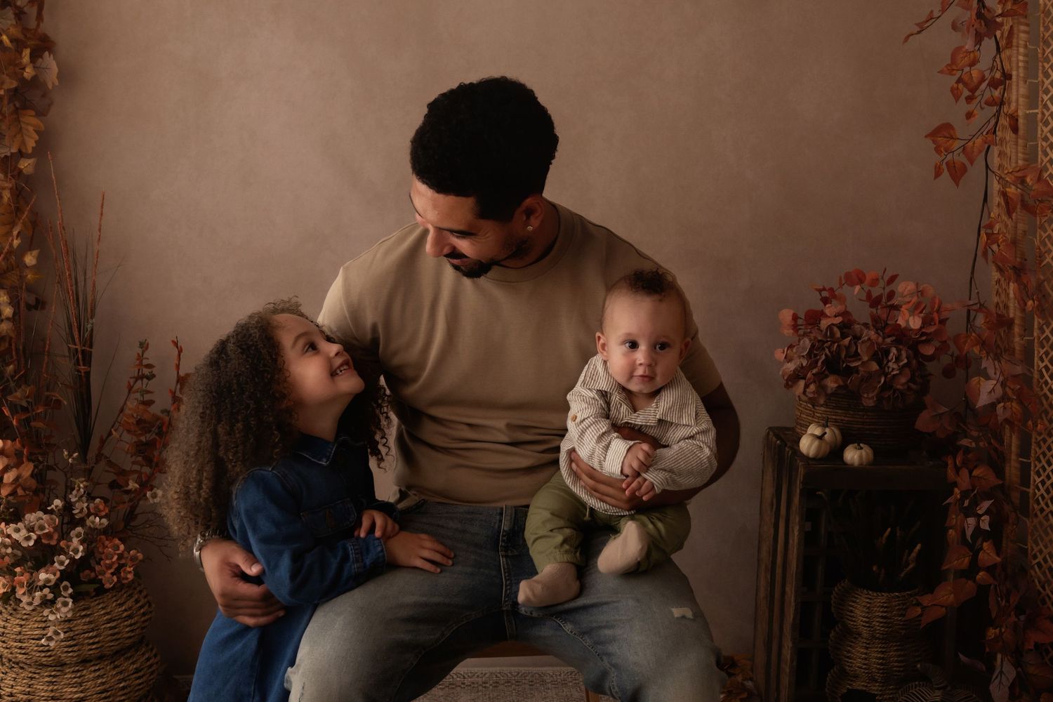 A father sits with his two young children, a toddler girl and baby boy, in a warm autumn-themed portrait studio.