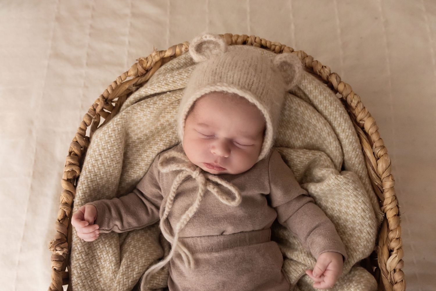 Sleeping newborn baby in bear bonnet hat and brown outfit nestled in wicker basket.
