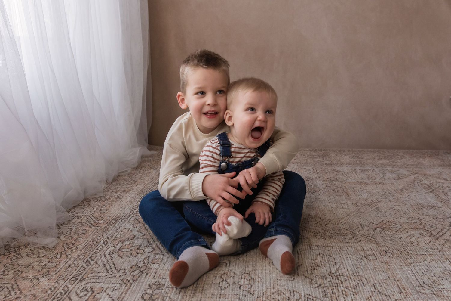 Two young brothers sit together on a rug, the older boy hugging the laughing baby sibling.
