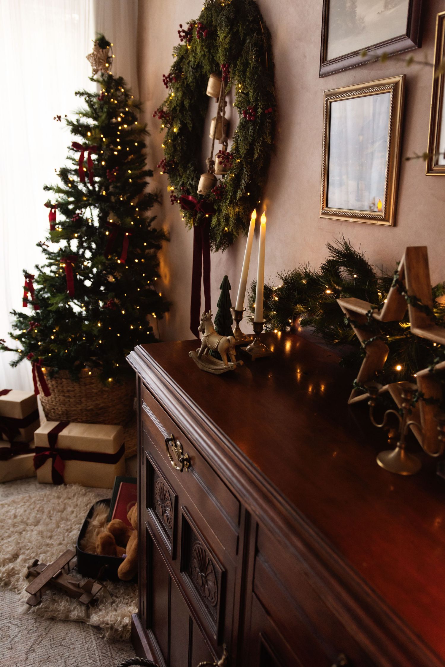 Cozy Christmas living room with decorated tree, gifts, wreath, candles, and garland on antique wooden dresser.