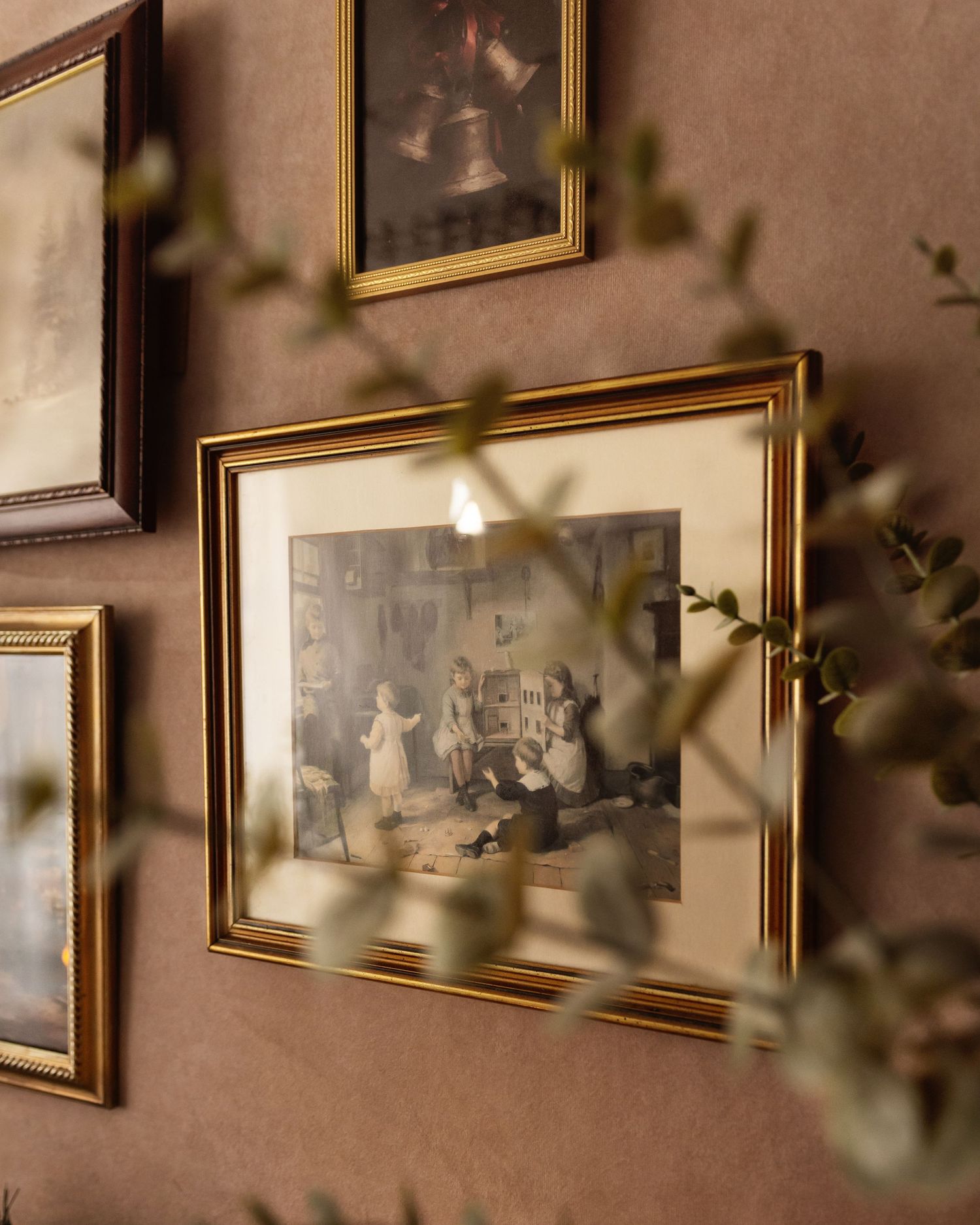 Gold-framed vintage artwork displayed on a dusty rose wall, surrounded by other antique paintings and soft green botanical branches.