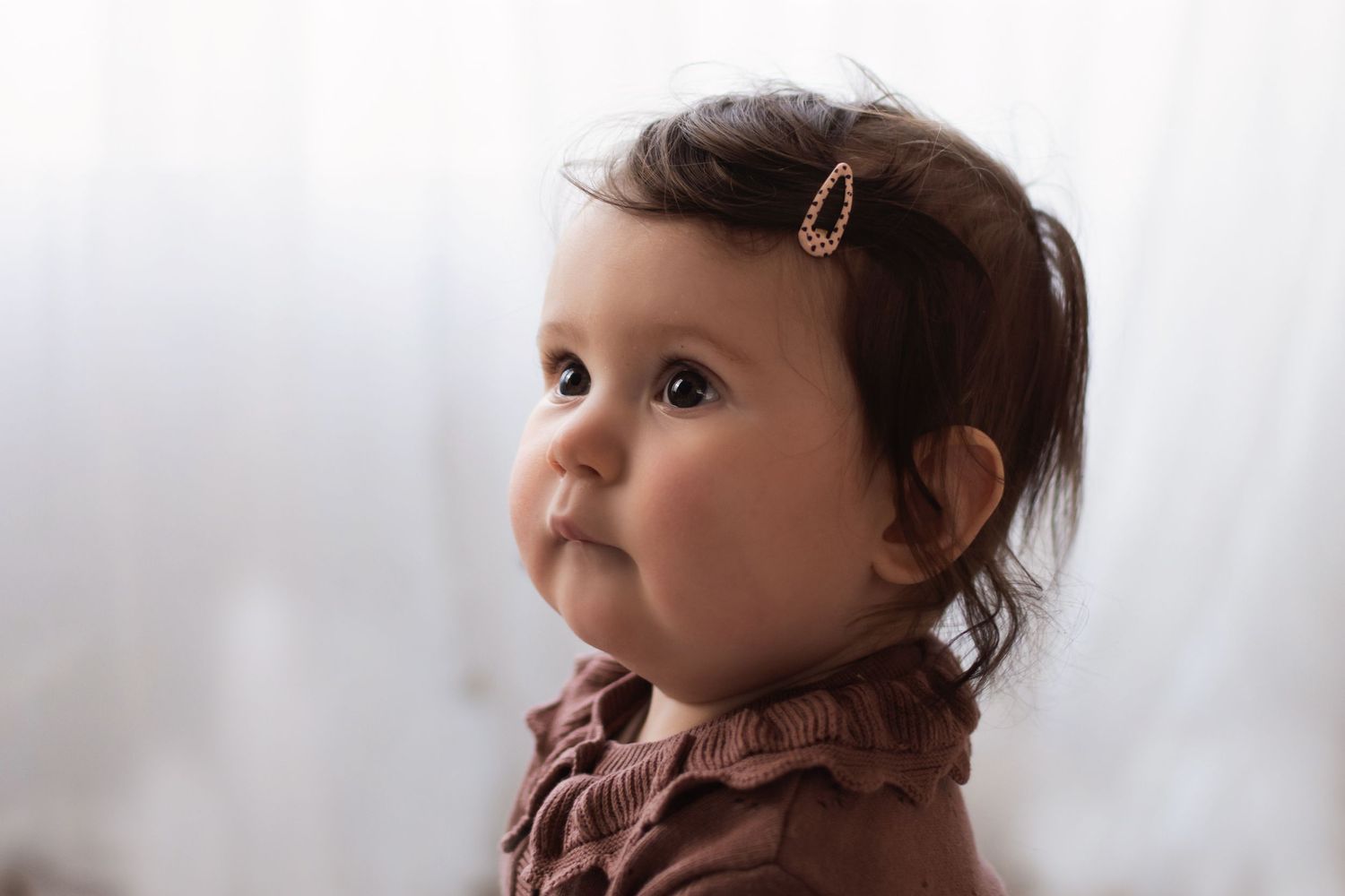 A sweet baby girl with a hair clip looks up thoughtfully against a soft white backdrop.