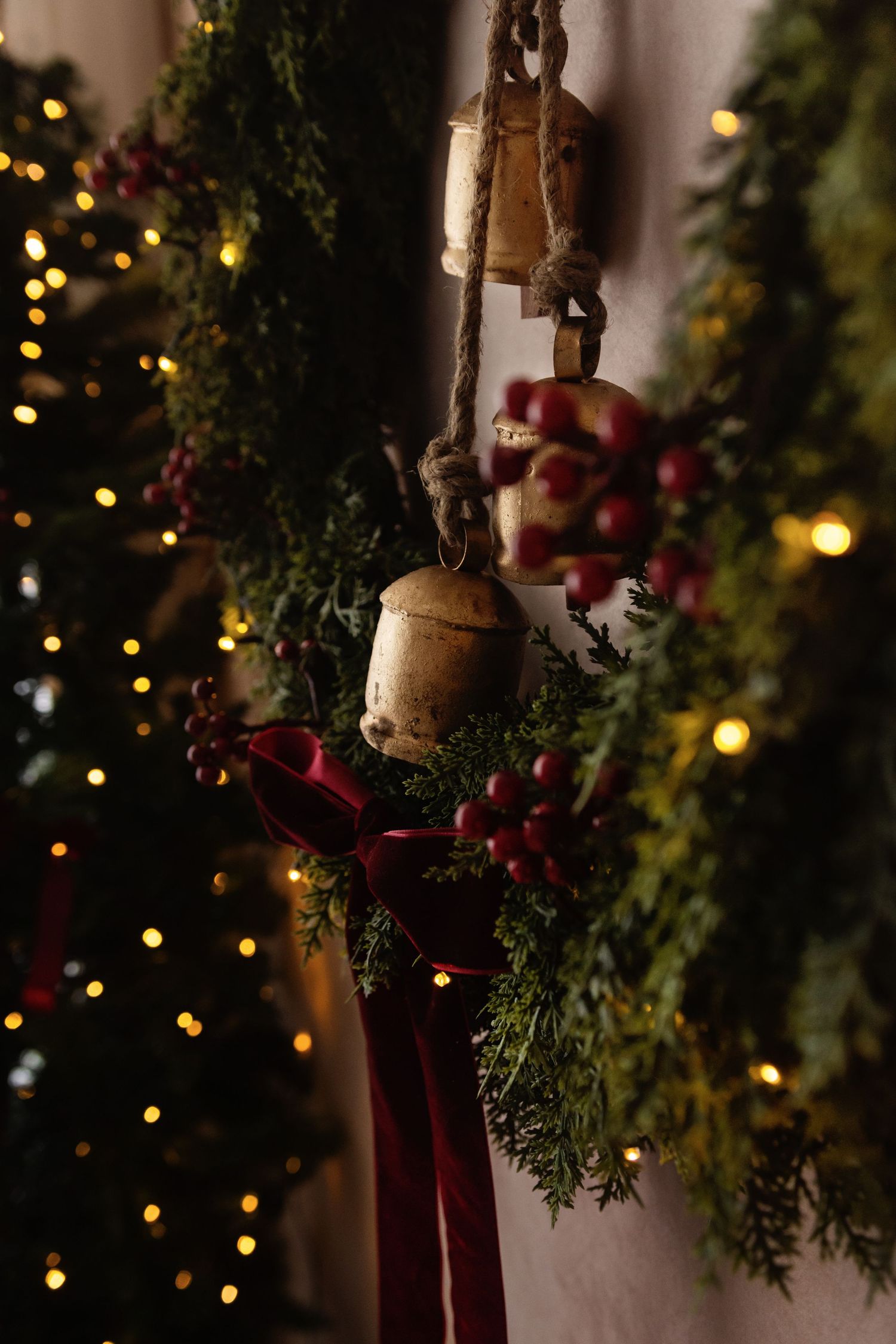 Rustic Christmas bells hanging with evergreen sprigs, red berries, and a crimson ribbon, with bokeh lights in background.
