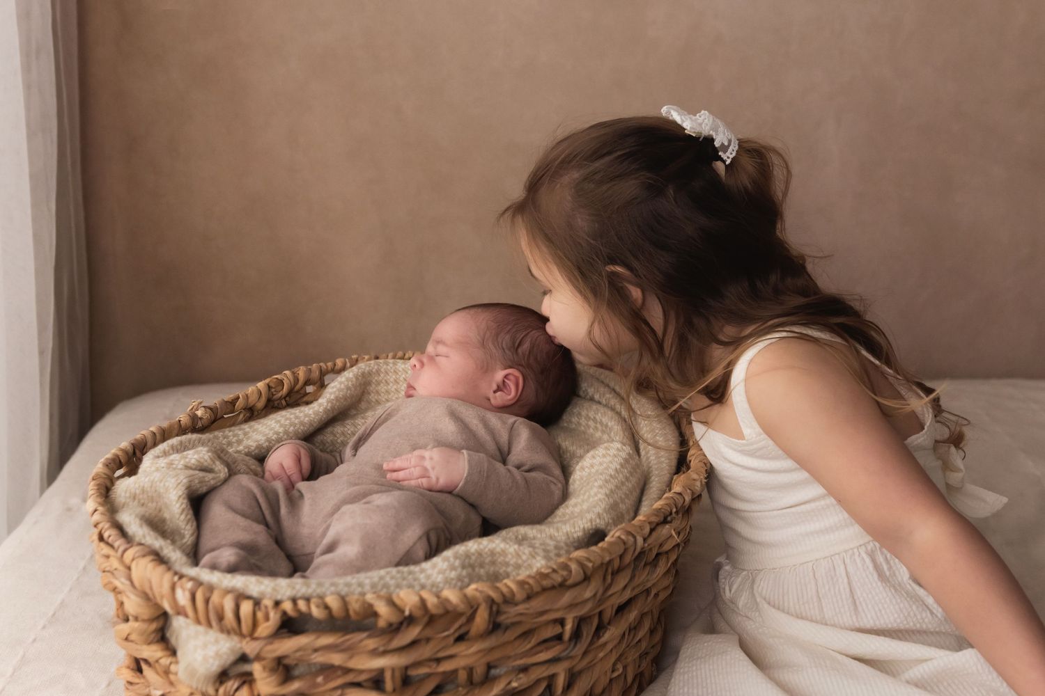 A young girl in a white dress lovingly kisses a sleeping newborn baby nestled in a woven basket.