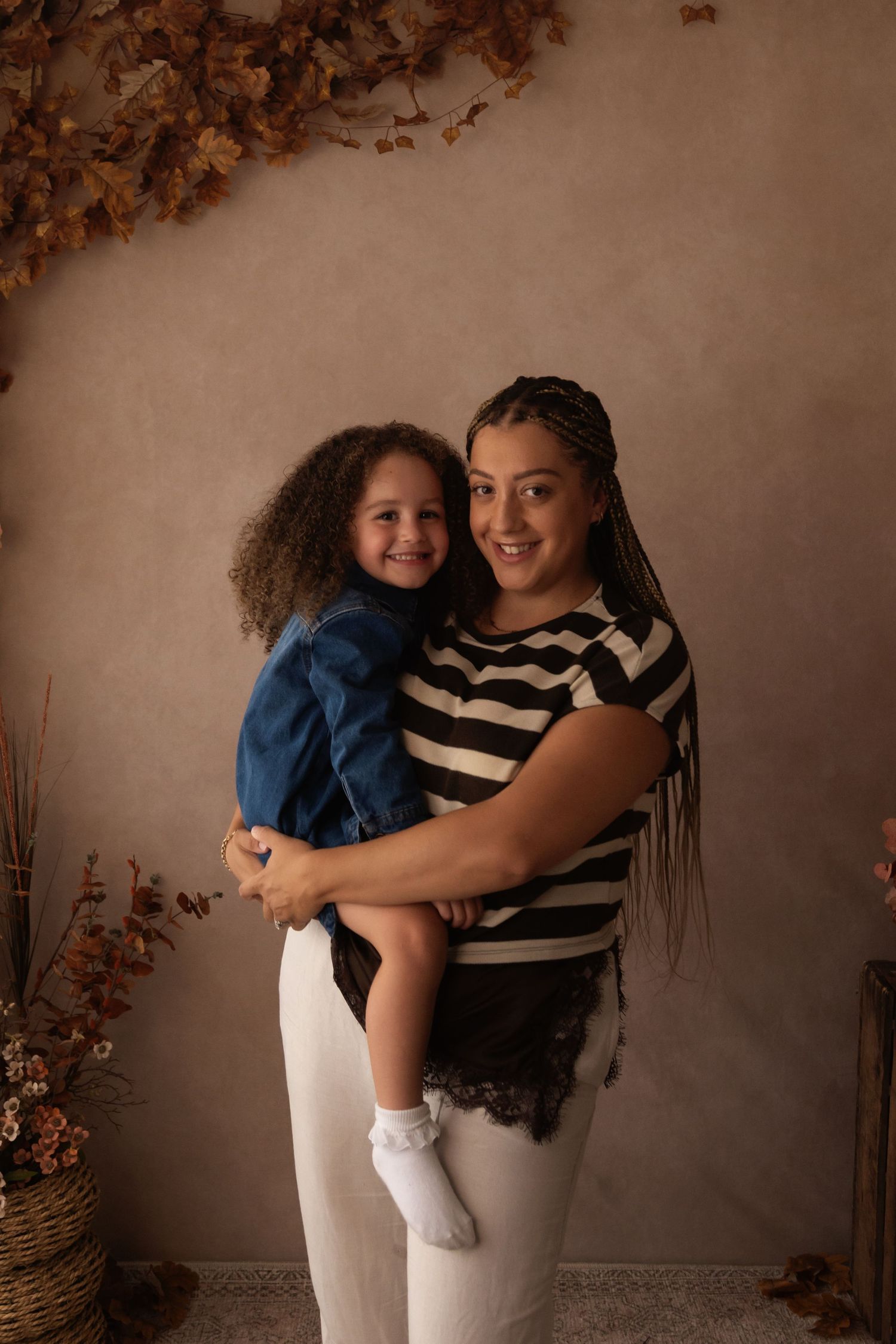 A woman in a striped shirt holds a young girl in a denim jacket, posing in a warm autumn-themed studio setting.