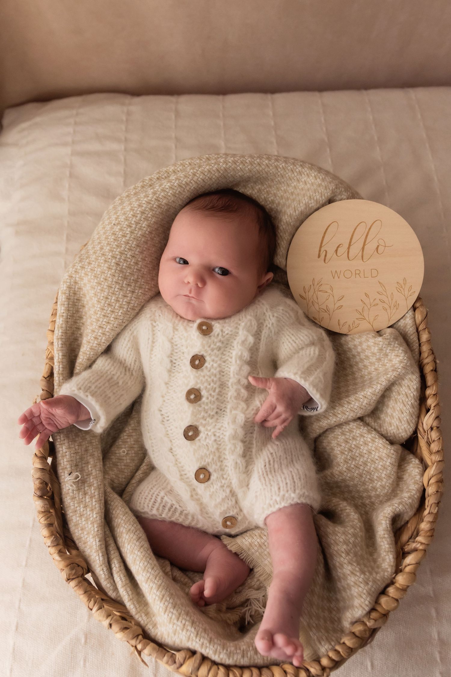 Newborn baby in cream knit romper lying in wicker basket with Hello World wooden sign.