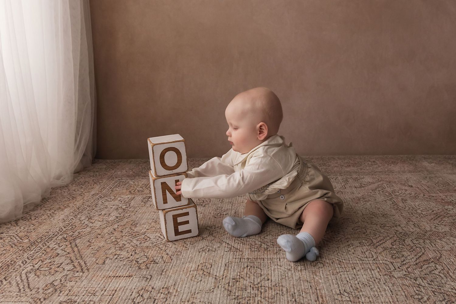 Baby in beige romper reaches for stacked wooden ONE blocks sitting on a patterned rug.